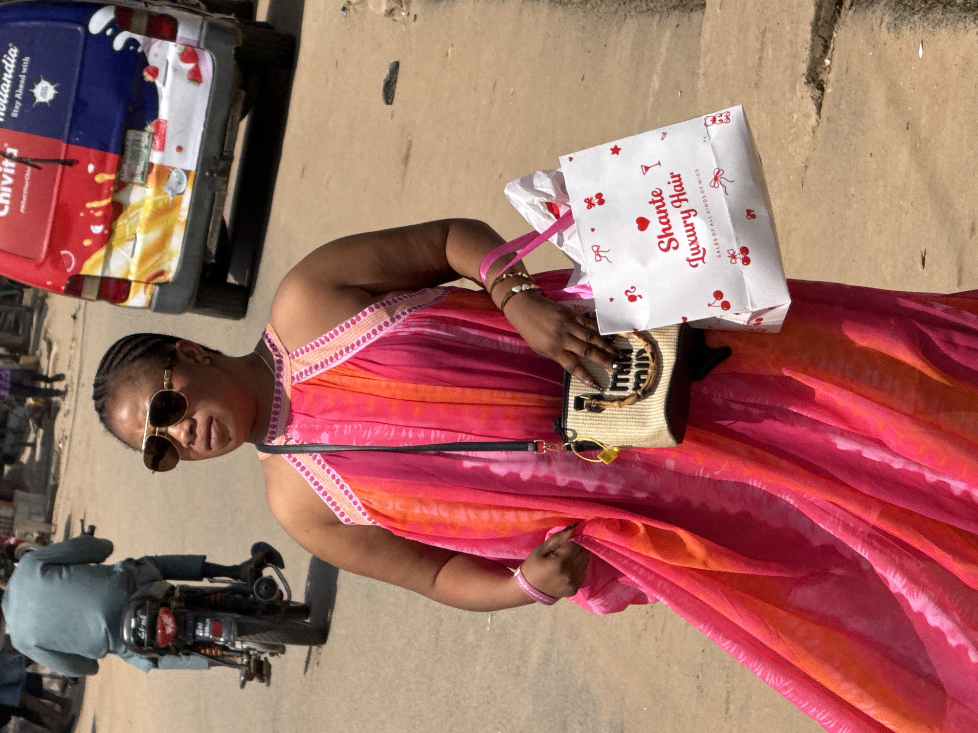 In Abuja, Nigeria, a woman in a flowing pink and orange tie-dye dress walks on a dusty street, holding a straw purse and a white shopping bag with red cherries and the words "Shante Luxury Hair" printed on it. She wears aviator sunglasses and a pink bracelet. Behind her, a person on a motorcycle in a blue tunic is visible, and a red and blue van with colorful decals is parked. The scene captures a moment of daily life on a sunlit urban street.