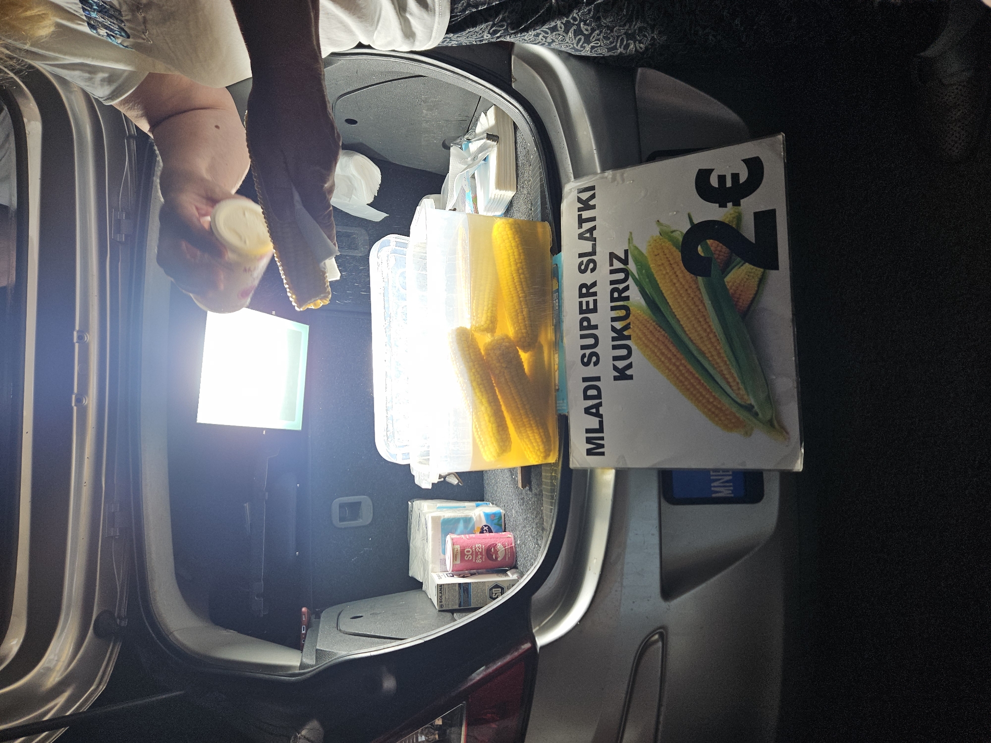 In the open trunk of a car, a person is preparing to season corn on the cob. Several ears of corn are submerged in a clear liquid within a plastic container. Next to the corn, there are stacks of paper napkins and other packaged goods. A sign in front of the trunk advertises "MLADI SUPER SLATKI KUKURUZ" (young super sweet corn) for "2€". The background is dark with a bright light illuminating the scene. The car's license plate, partially visible, reads "MNE". The overall setting appears to be a roadside stall or a temporary setup for selling food.