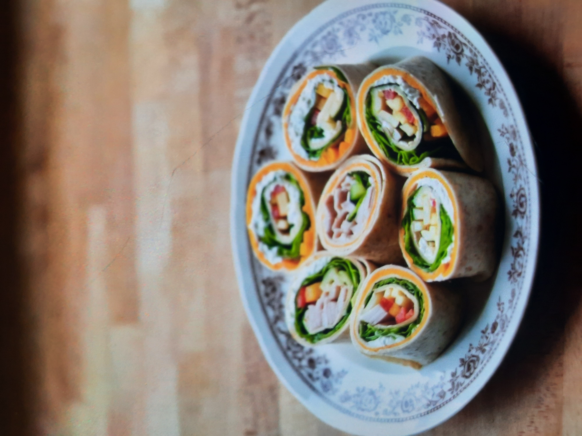 Seven pinwheel wraps are arranged on a white plate with a dark brown decorative border, set on a light brown wooden surface. The wraps are made with tortillas and filled with various ingredients, including green lettuce, diced red and yellow bell peppers, and possibly cheese or turkey. The plate is positioned slightly off-center to the right. The wooden surface appears to be a table or countertop. No people are present in the image. The lighting suggests it is indoors. The image does not contain any information that would allow for the determination of its location in Rosenheim, Germany.