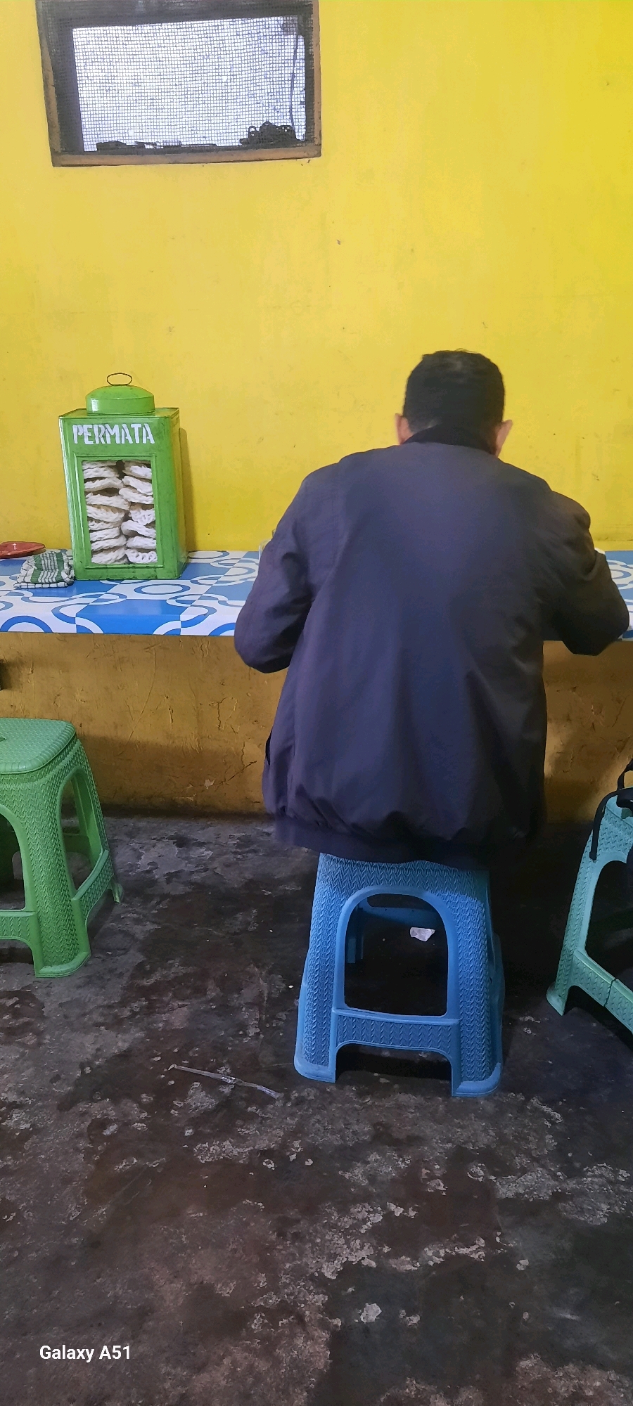 A rear view of a man wearing a dark jacket sits on a blue plastic stool in front of a table covered with a blue and white patterned cloth. The table holds a green tin container labeled "PERMATA" filled with snacks and a folded green and white striped cloth. To the left of the table, a green plastic stool is visible. The background features a yellow wall with a small window covered by a mesh screen. The floor is a dark, textured surface. The scene appears to be an indoor setting, possibly a local eatery or shop in Selopuro, Indonesia.