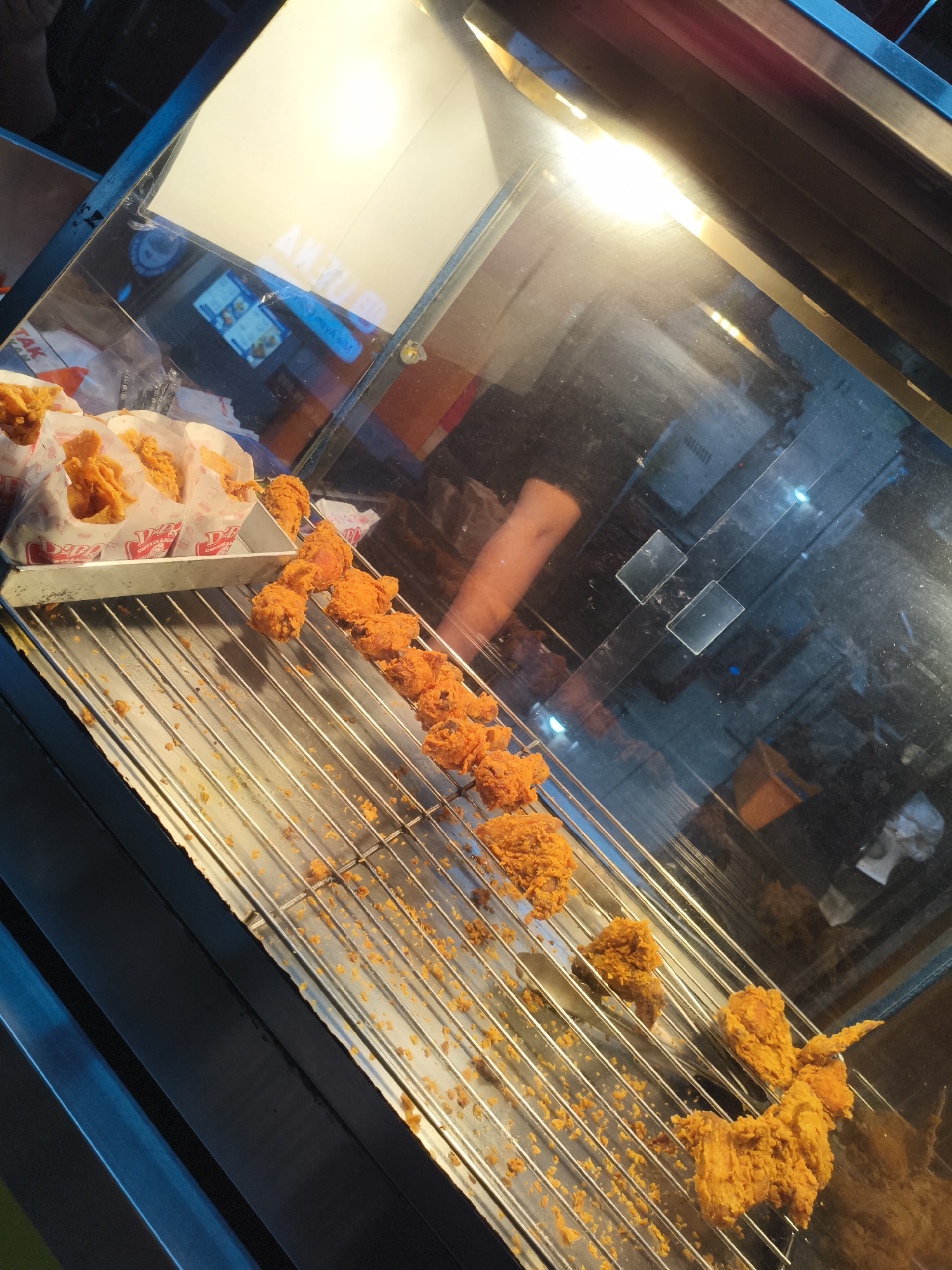 A high-angle shot depicts a heated display case filled with fried chicken. The case contains a tray of chicken pieces wrapped in branded paper, alongside several chicken pieces arranged on a metal wire rack. Crumbs and small fragments of fried chicken are scattered along the rack's surface. The lighting inside the case is bright, creating reflections on the glass surfaces. A person's arm is visible within the reflected background. This image was taken in Depok, Indonesia.