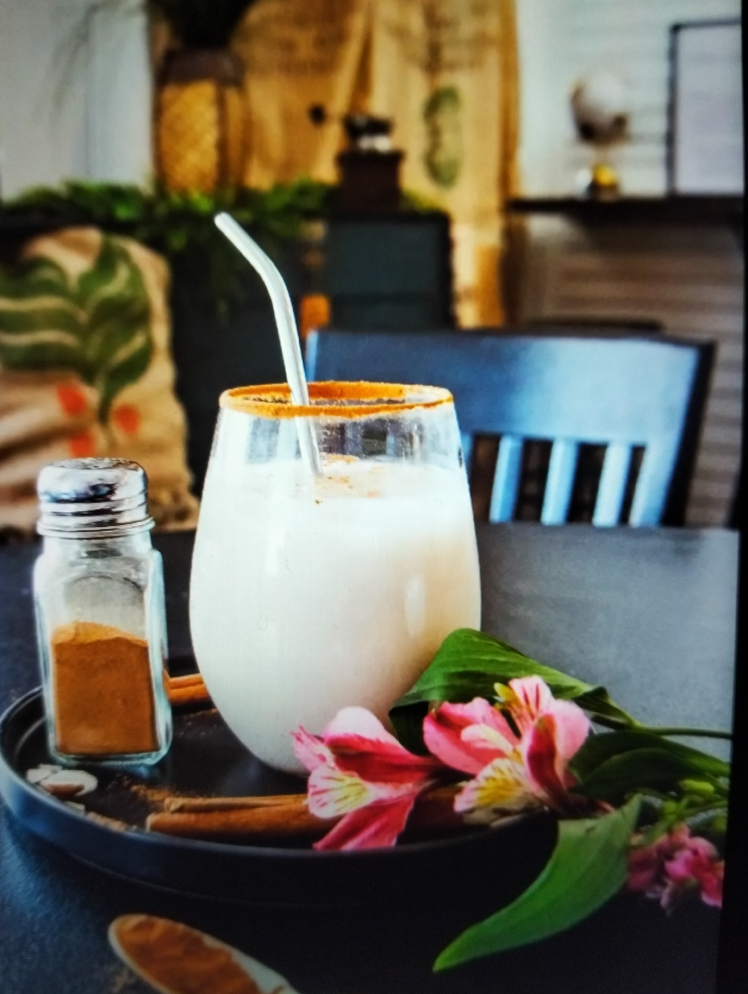 An eye-level, medium shot captures a glass of creamy beverage with a cinnamon-dusted rim, a white straw, and a small glass shaker filled with cinnamon powder, all arranged on a dark, circular tray. Two cinnamon sticks and a cluster of pink and yellow flowers with green leaves are also on the tray, positioned near the glass. The background is blurred, showing hints of furniture, greenery, and patterned fabric. The image does not provide any information about the city of Payagaboh, Indonesia.