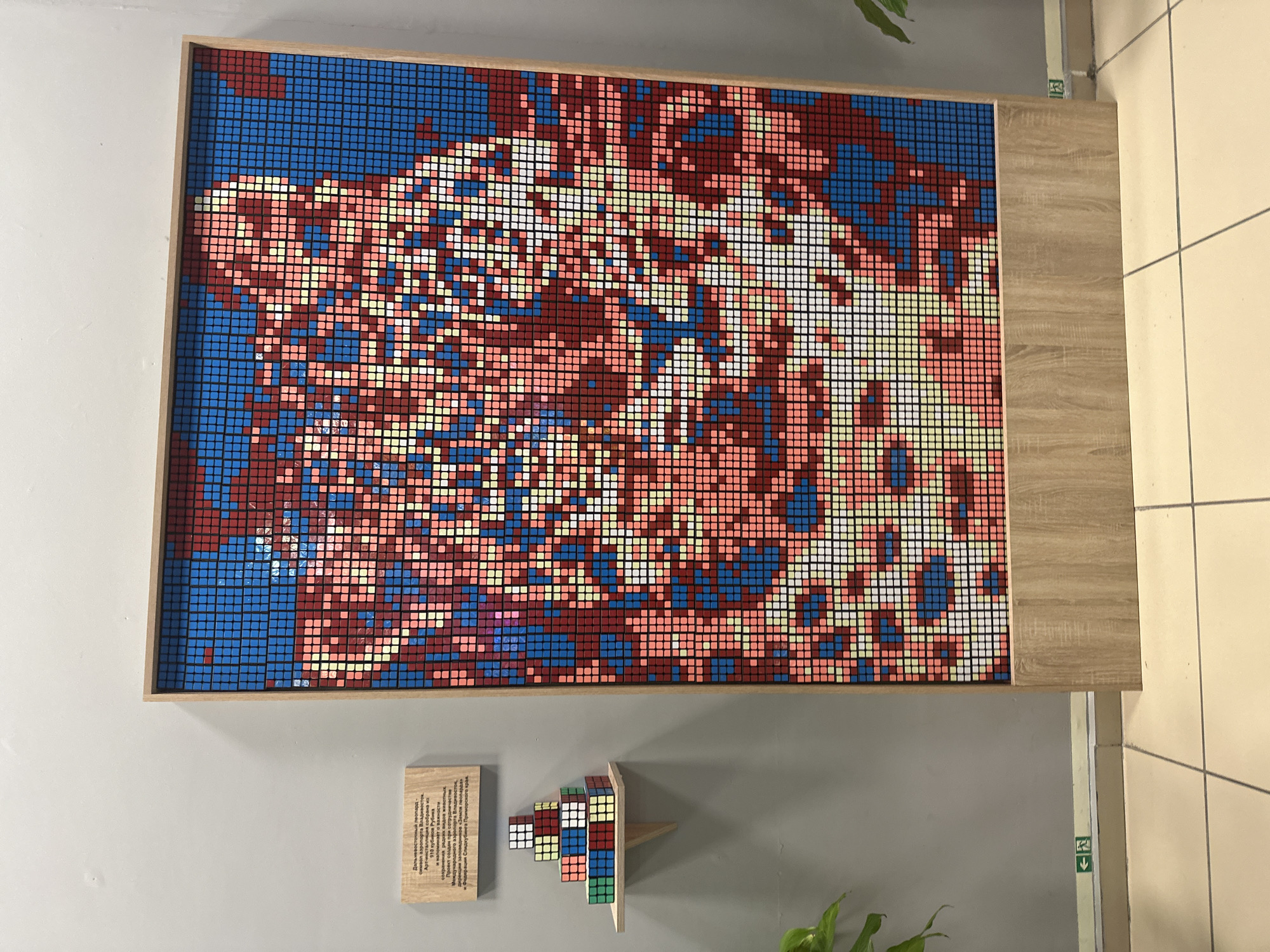 The image shows a large mosaic artwork composed of many small Rubik's cubes, arranged on a wall. The mosaic itself depicts an abstract pattern with various shades of blue, red, pink, yellow, and white. The cubes are arranged in a grid, forming a cohesive image within a light-colored wooden frame. Below the large mosaic, there are two smaller items displayed on the wall: a wooden plaque with text and a small, tiered sculpture made of Rubik's cubes. The sculpture is colorful and seems to be a staircase-like structure. The overall setting appears to be an indoor exhibition space or gallery, with a plain grey wall and a section of a wooden surface to the right, possibly part of a display table or furniture. The lighting suggests an indoor environment, possibly during the daytime.
The wooden plaque has text written in Cyrillic script. The text appears to describe the artwork or its context, possibly mentioning the location "Artyom, Russia" and details about the project or artist. The text is too small to be fully deciphered from the image, but it is a notable detail of the display. The presence of a plant in the lower right corner suggests a decorative element in the space. The overall impression is of an art installation that utilizes a popular toy, the Rubik's cube, to create a visually interesting mosaic and accompanying sculptures.