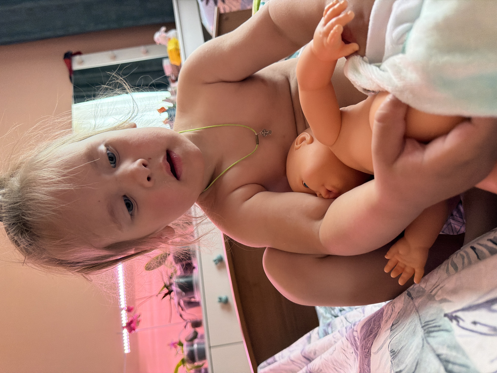 A young child, likely a toddler, with light blonde hair and blue eyes, is captured in a close-up, seemingly lying down and looking upwards towards the camera with an open-mouthed expression. The child is shirtless, wearing a thin green necklace with a small, metallic pendant. They are holding a light-skinned baby doll, which is partially covered by a white and light blue patterned fabric, possibly a blanket or diaper. A hand, possibly an adult's, is also visible near the doll, assisting or playing alongside the child.
The setting appears to be indoors, likely a bedroom or play area in a home in Chaikovsky, Russia. In the background, a mirror reflects parts of a room, and to the left, several plants are visible under a strip of vibrant pink grow lights, suggesting a dedicated area for horticulture. A patterned bedspread or blanket is visible in the lower right portion of the image. The overall mood is one of innocent playfulness. No visible text is present in the image.