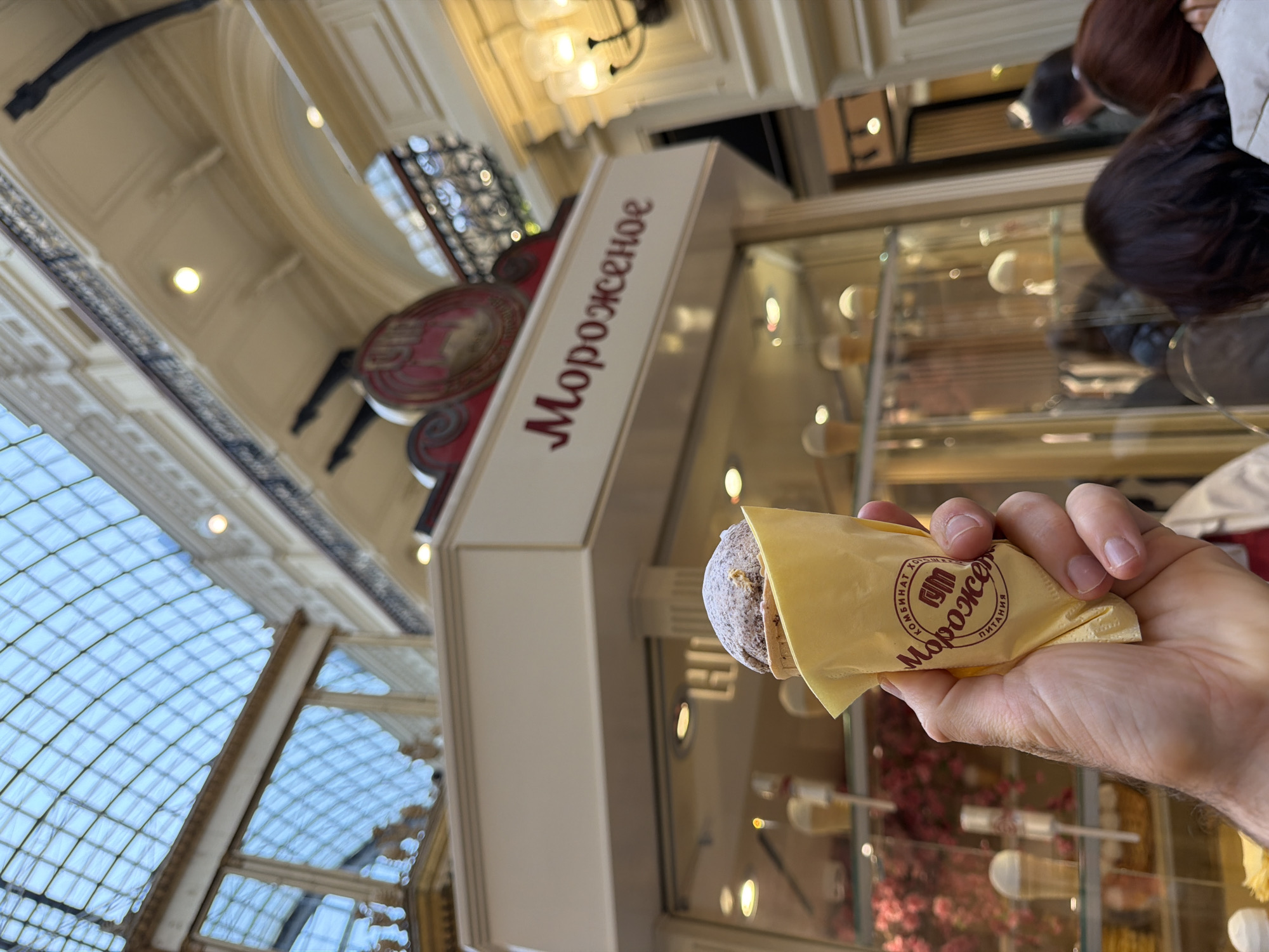 A person holds an ice cream cone in front of a kiosk in GUM department store, Moscow, Russia. The ice cream is wrapped in a yellow paper wrapper printed with the word “Мороженое” (ice cream) in red. The kiosk also has the word “Мороженое” in red. Behind the kiosk are glass display cases. The architecture of GUM is visible, including the arched ceiling and ornate decorations. Other people are present in the scene.