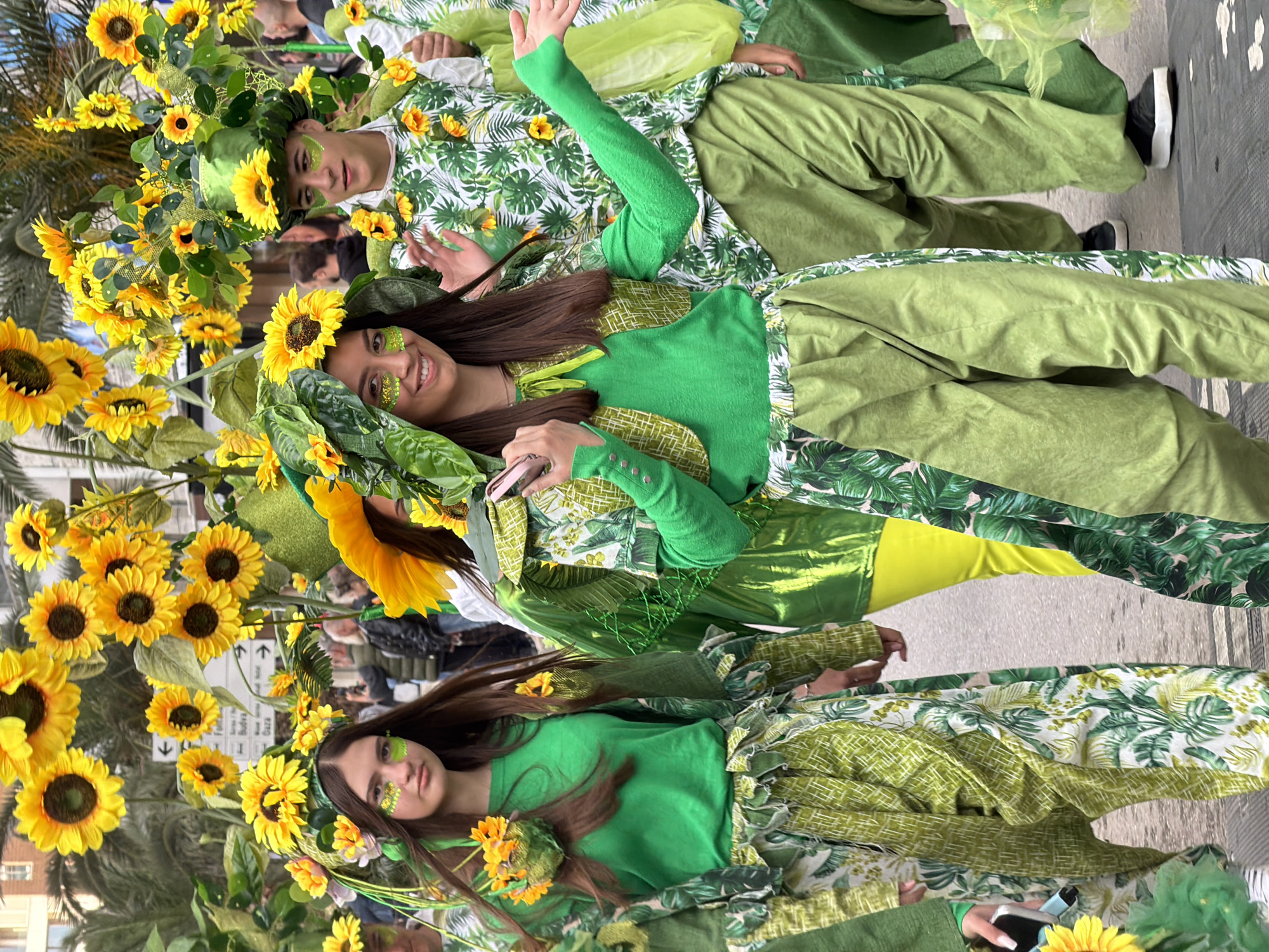 The image shows three people in green and yellow costumes with sunflower decorations, likely participating in a festival or parade. The person in the middle is smiling and waving, holding a mobile phone. The background appears to be a street in Budva, Montenegro, with other people and buildings visible in the distance. Road signs are visible in the background, in front of palm trees. The costumes appear to be inspired by nature.