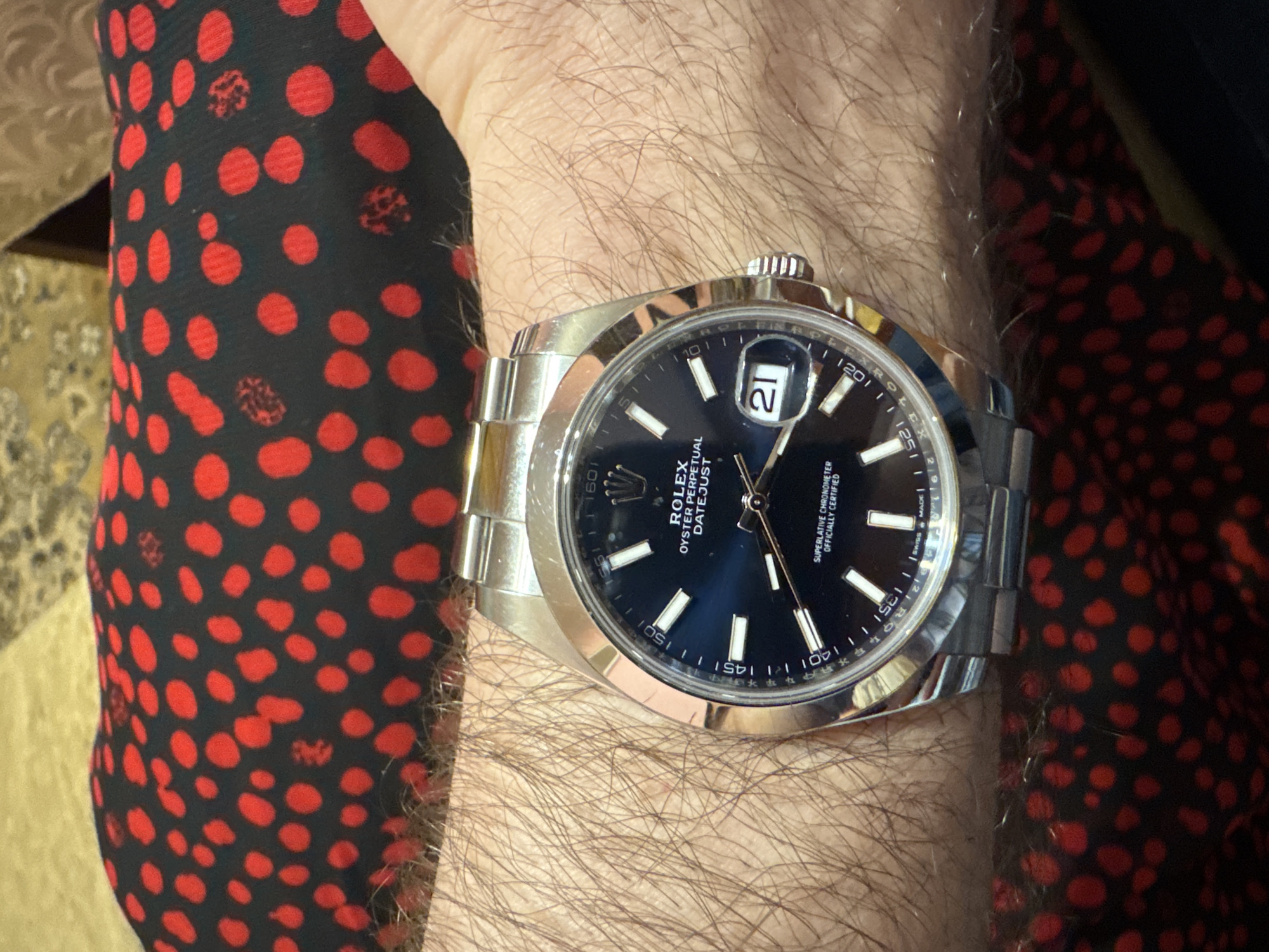 The image shows a person's wrist with a silver Rolex Datejust watch. The watch has a black dial with white hour markers and a date window at 3 o'clock showing the number 21. The watchband is silver. The person's arm is hairy. The person is wearing a garment with a black background and red polka dots. There is no location in Plovdiv, Bulgaria visible in this image.