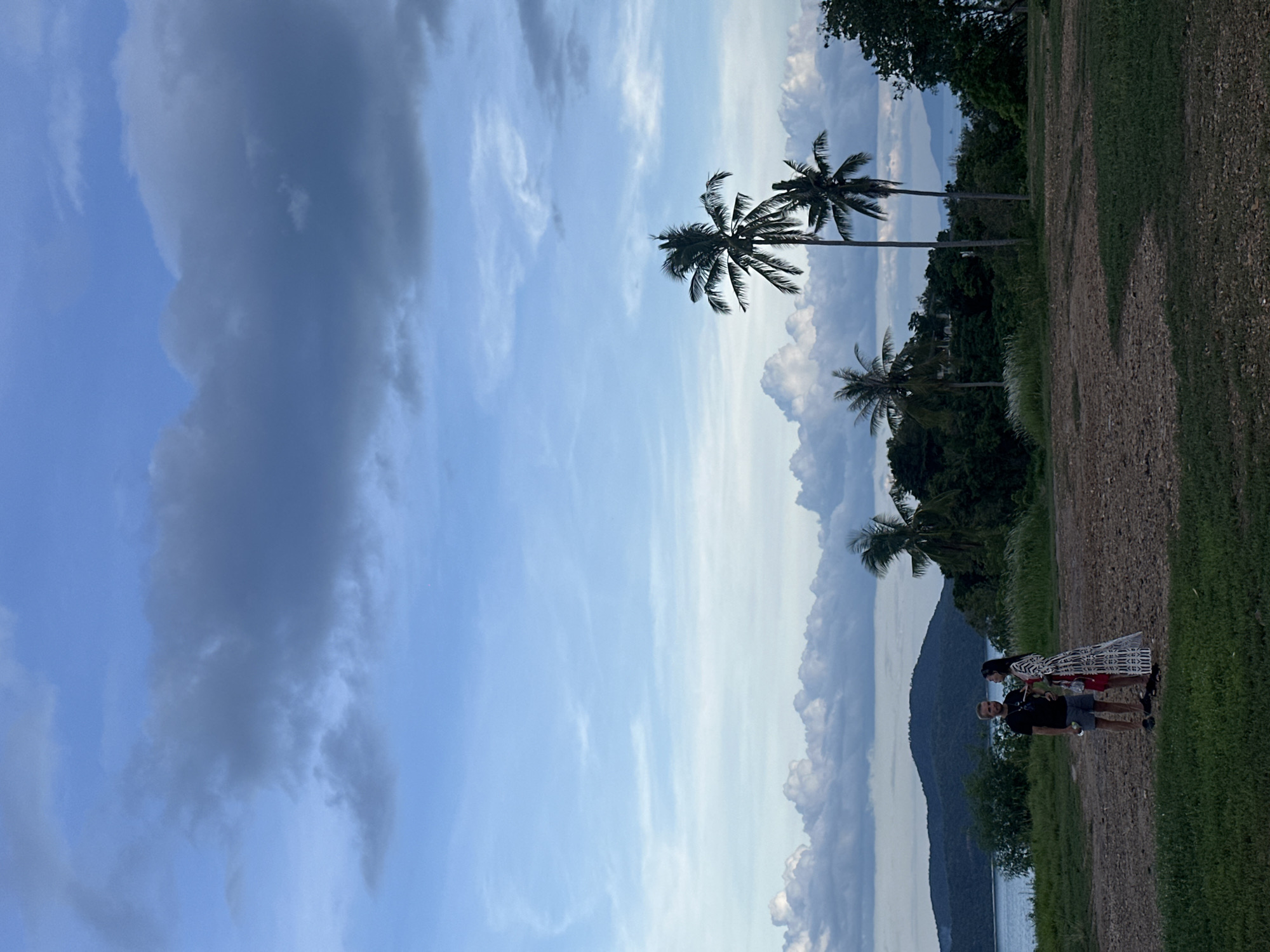 This image captures an outdoor tropical scene in Baan Pang Ka, Thailand, under an overcast sky. In the mid-ground, two individuals are standing on an unpaved area consisting of light brown earth and patches of dark green grass. The male, positioned on the left, wears a dark t-shirt, light shorts, and dark sandals, holding a white bottle in his left hand. The female, to his right, wears a long, patterned black and white dress and holds a red object, possibly a small bag, in her left hand. Both individuals are facing each other, suggesting a conversation or interaction.
The background features a large body of water, likely the sea, which separates the foreground from a distant, forested landmass with undulating hills. Lush green vegetation, including numerous trees and shrubs, covers the landscape. Several tall palm trees are prominent; one stands directly behind the female, and two more are visible further to the right. The sky overhead is largely covered by clouds, ranging from light blue to dark grey and white cumuliform formations, indicating either late afternoon or early evening lighting conditions.