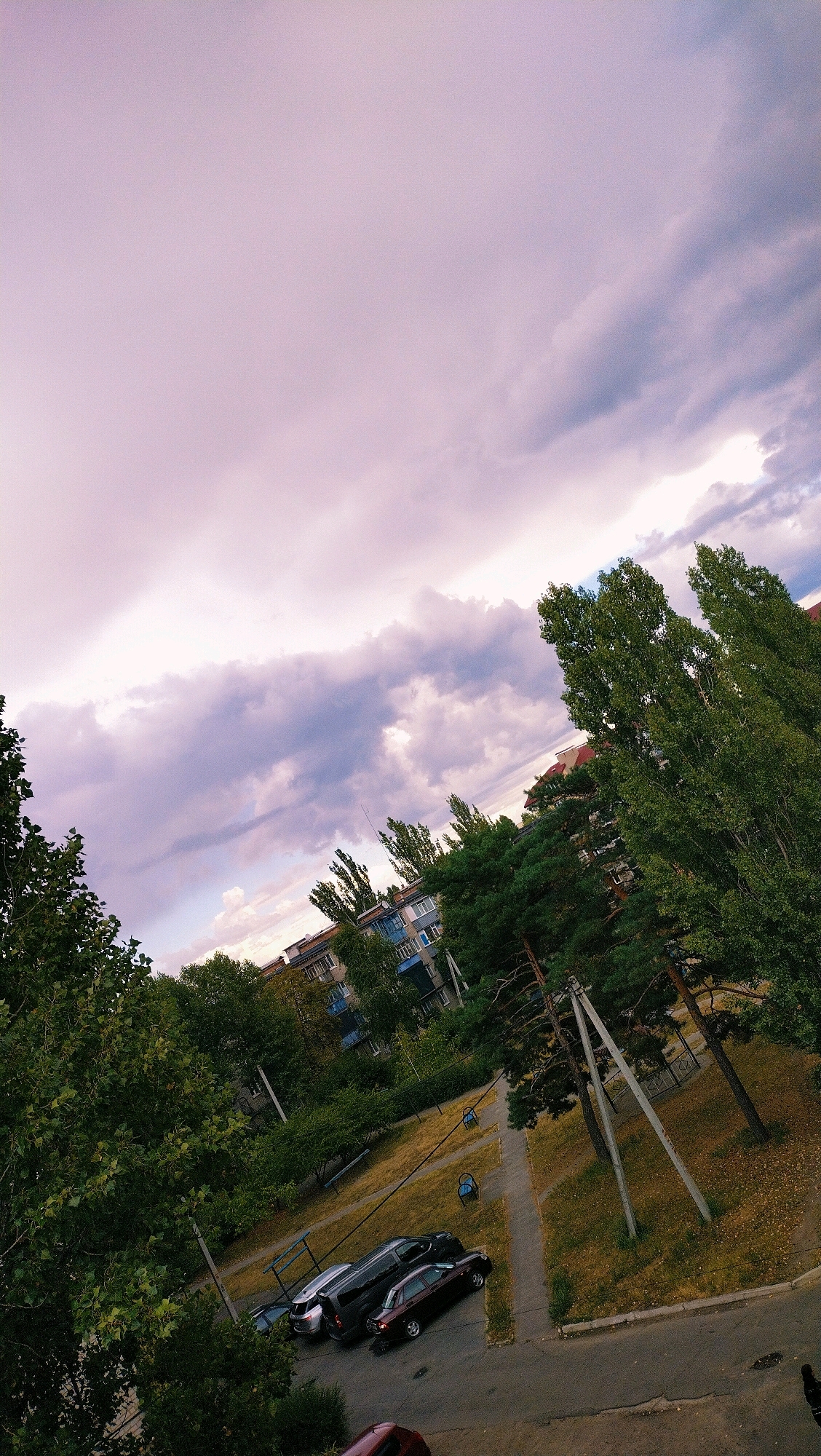 The image captures a view of an apartment building courtyard in Horishni Plavni, Ukraine, under a cloudy sky. The sky is predominantly filled with light purple and gray clouds, with hints of brighter sky visible. In the foreground, lush green trees frame the scene, with some appearing to be deciduous and others coniferous.
In the middle ground, an apartment building with a light blue facade and white window frames is visible, surrounded by more trees. The courtyard area features a paved walkway and a patch of dry grass. Several parked cars are visible on the asphalt and grass area. These include a dark-colored sedan, a silver sedan, and a dark gray van.
There are no people visible in the image. The environment appears to be a typical residential area with trees and apartment buildings. The lighting suggests it is either early morning or late afternoon, given the soft, diffused light.