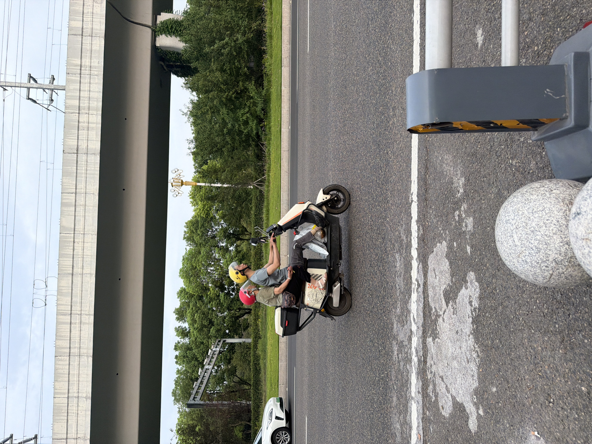 In the city of Jiangjin, China, two individuals are riding on a white electric scooter on a multi-lane paved road. The rider in the front wears a yellow helmet and a grey t-shirt. The passenger behind, possibly an older woman, wears a red helmet and a green t-shirt. Both are facing forward, and the passenger has her arms around the rider. The scooter has a black storage box mounted on the rear. The road has white lane markings, and on the left side, a white car is visible. In the background, there is a dense line of green trees, and above that, a large elevated concrete overpass with power lines and utility poles. The sky is overcast. In the foreground, the asphalt road surface shows patches of lighter grey material, possibly from a previous spill or repair. A grey traffic bollard with yellow and black hazard markings is partially visible on the right, and a round grey stone object is in the lower left.