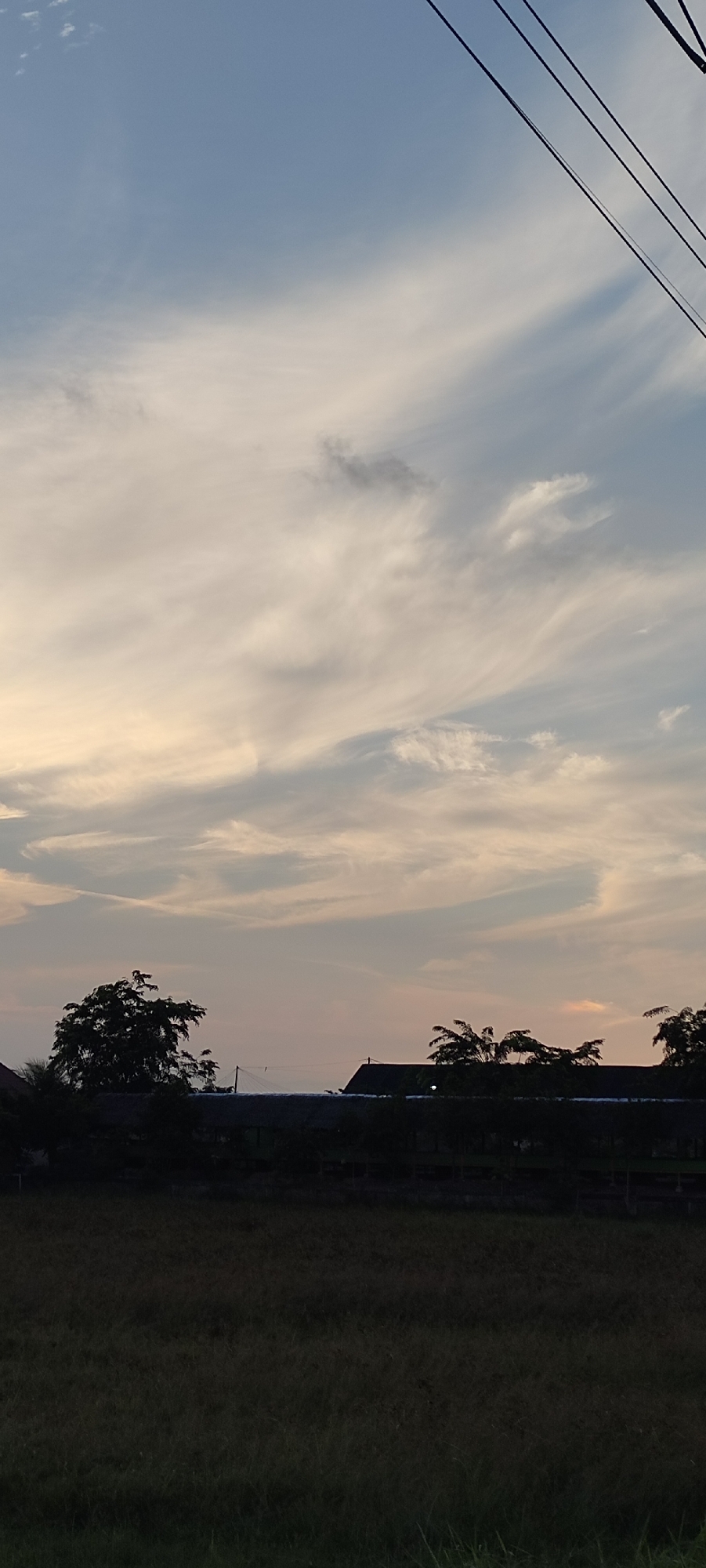 The image depicts a scene in Cotbabadatunong, Indonesia, at dusk. The sky is a soft blend of blue, white, and peach hues, with wispy clouds stretching across it. Silhouetted against the fading light are trees and the roofs of several buildings. In the foreground, a dark field of grass, likely a rice paddy, stretches towards the horizon. Three dark, taut power lines are visible against the sky in the upper right corner of the image.