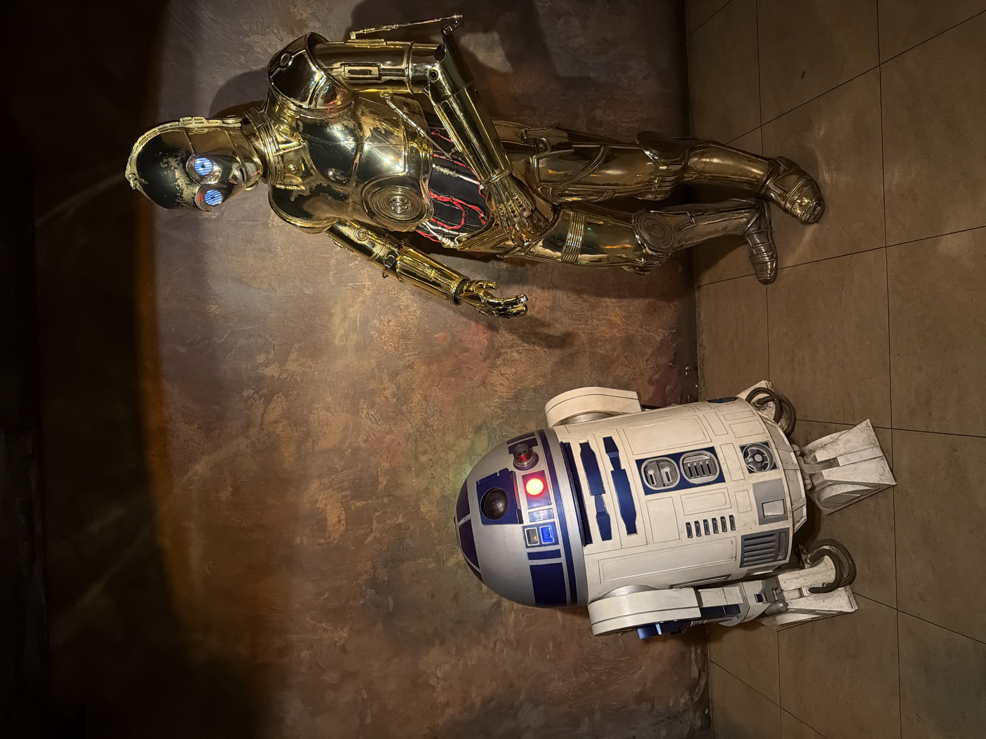 A high-angle shot depicts static figures of C-3PO and R2-D2 against a textured, mottled brown backdrop, resembling a wall or floor. C-3PO, a golden humanoid robot, lies on its side, with visible internal wiring and metallic limbs. R2-D2, a white and blue cylindrical droid with various panels and attachments, sits upright near C-3PO. The location is not discernible from the provided image.