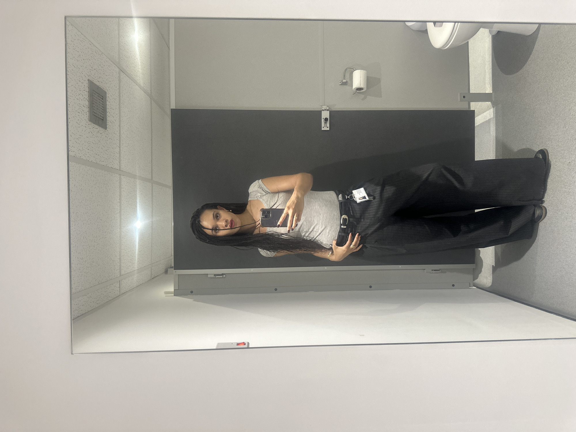 A mirrored image of a woman taking a selfie in a bathroom is captured. The bathroom features a white tiled ceiling with recessed lighting, a grey wall, a black partition, a toilet, and a white tiled floor. The woman has long, braided hair and is wearing a light grey t-shirt and dark, wide-legged trousers with a belt. She is holding a smartphone, presumably taking the selfie. The location is not specified, and there is no information indicating that it is in the city of London, United Kingdom.