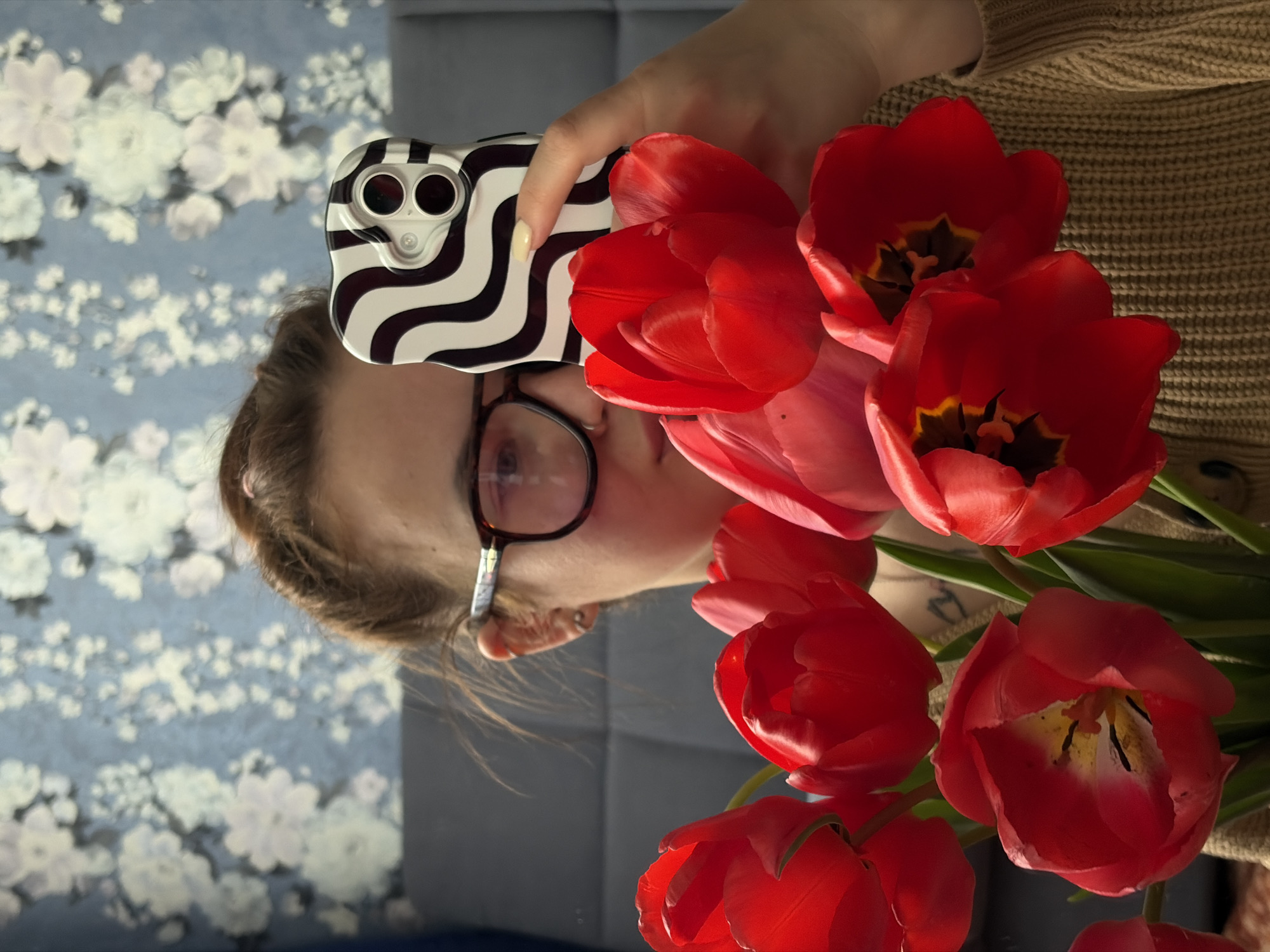 The image captures a person indoors, holding a smartphone and a bouquet of red tulips. The person, seen from the side profile, has light brown hair pulled back, wears dark-framed glasses, and is dressed in a light brown knitted sweater or cardigan. Their right hand holds a smartphone encased in a distinctive black and white wavy-striped cover, with the dual camera lenses visible on the back.
Prominently featured in the foreground is a vibrant bouquet of red tulips with green stems. Several tulips are fully bloomed, revealing their yellow and dark centers, while others remain partially closed, showcasing their rich red petals. The person appears to be interacting with the flowers, potentially admiring them or preparing to take a photograph, as the phone is held in a viewing/capturing position.
The background consists of a light blue wall adorned with a delicate white floral wallpaper pattern. Behind the person's head and shoulder, a grey upholstered surface, possibly a couch or a headboard, is visible. The lighting within the scene suggests it is daytime. There is no visible text in the image. The setting is identified as Novopokrovskaya, Russia.