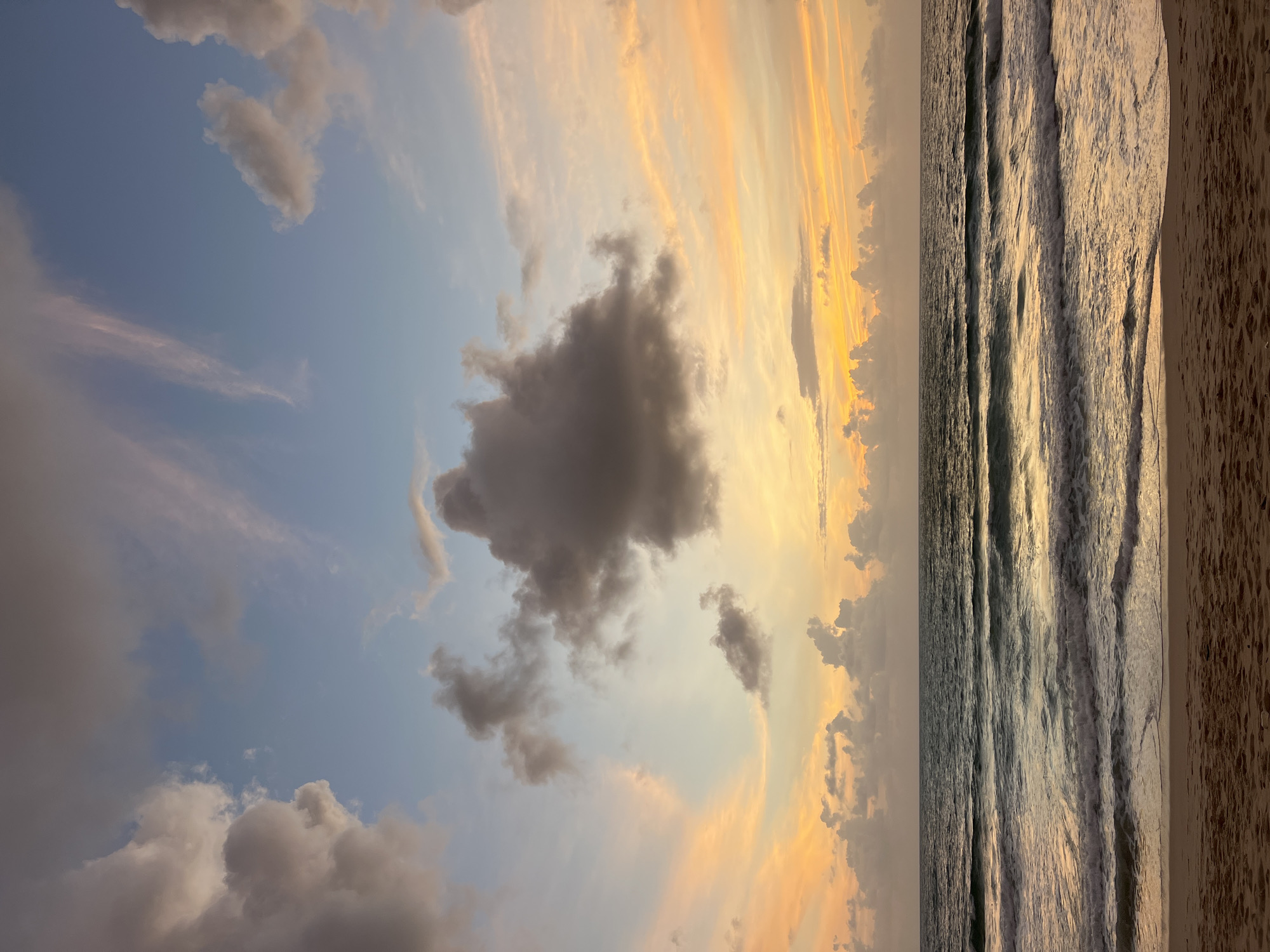 The image depicts a serene beach scene at sunset in Choeng Thale, Thailand. The sky is a canvas of colors, with soft oranges and yellows blending into hues of blue and grey as clouds drift across. The ocean is calm, with gentle waves rolling onto the sandy shore. The sand itself is a light brown, showing subtle textures from the tide. There are no people or man-made objects visible in the frame, creating a natural and tranquil atmosphere. The lighting suggests the golden hour, casting a warm glow over the entire landscape.