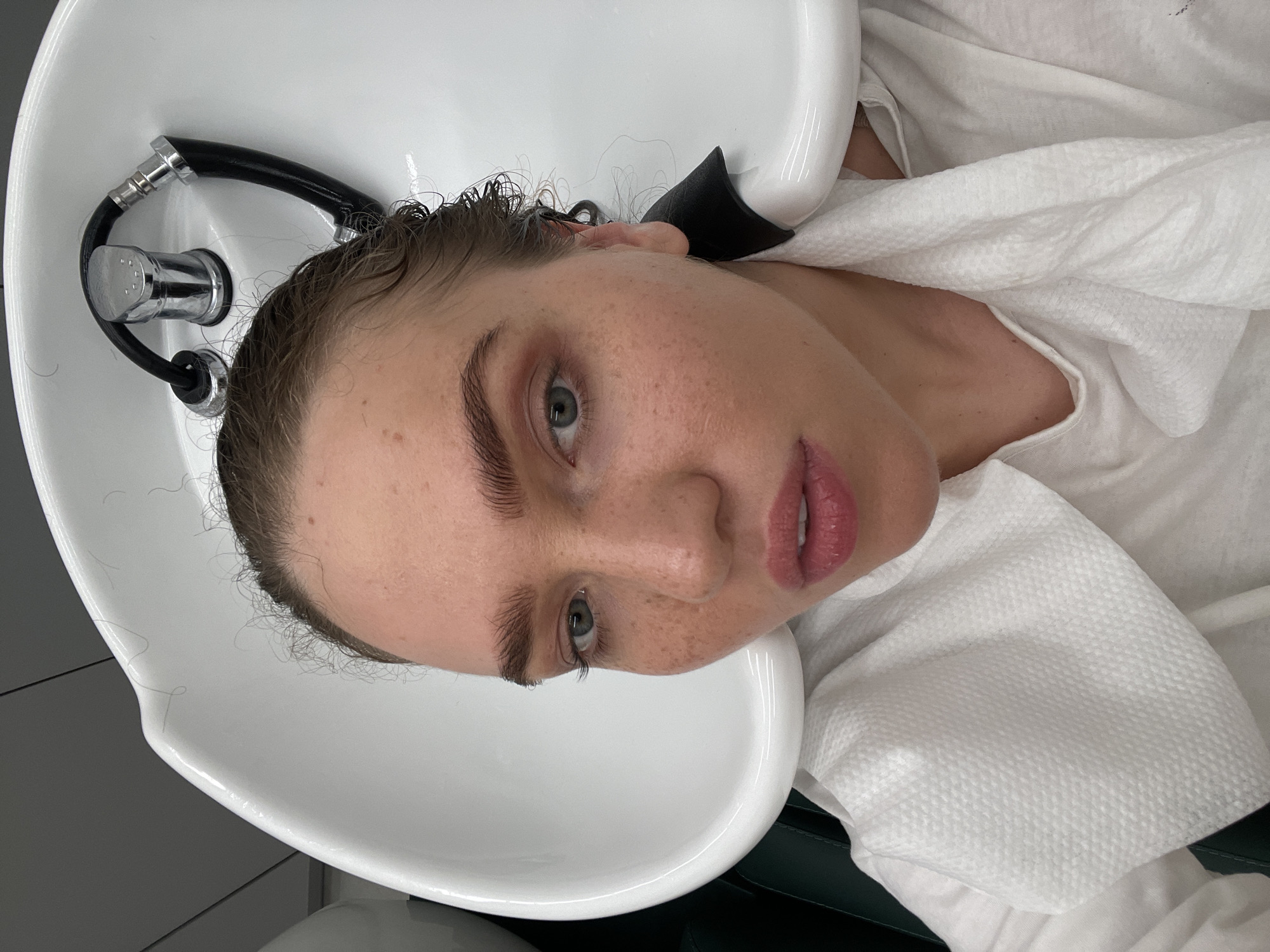 A woman is reclining in a white salon hair washing sink. She has fair skin, light brown hair that is slightly damp, and light-colored eyes. The sink is positioned next to a chrome faucet with a black hose. The woman is wearing a white towel draped around her shoulders. The background is indistinct, but suggests a hair salon setting. The image does not depict any specific location in Moscow, Russia or any other city.
