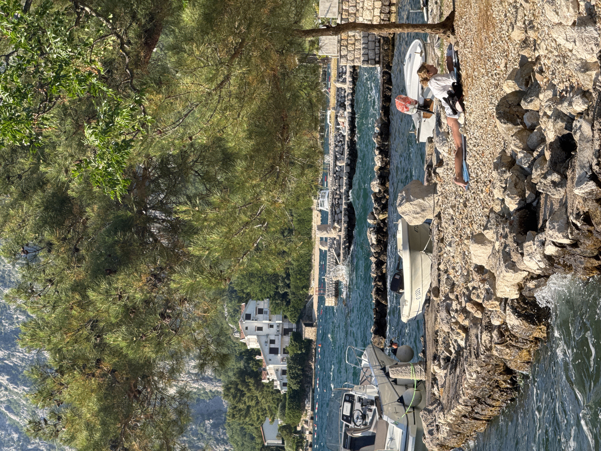The scene is a bright, sunny day on the coast of Dobrota, Montenegro. The landscape includes a stone embankment and a rocky shoreline. A body of water, likely the Adriatic Sea, stretches into the distance.
A woman in a white top and dark bottoms stands on the embankment near a white inflatable boat, appearing to be interacting with it. There's a possible red object in her left hand, and she has a black bag or backpack.
Several boats of varying sizes are moored in the water. In the background, there are buildings, trees, and a hint of mountainous terrain. A large tree frames the scene on the left side.