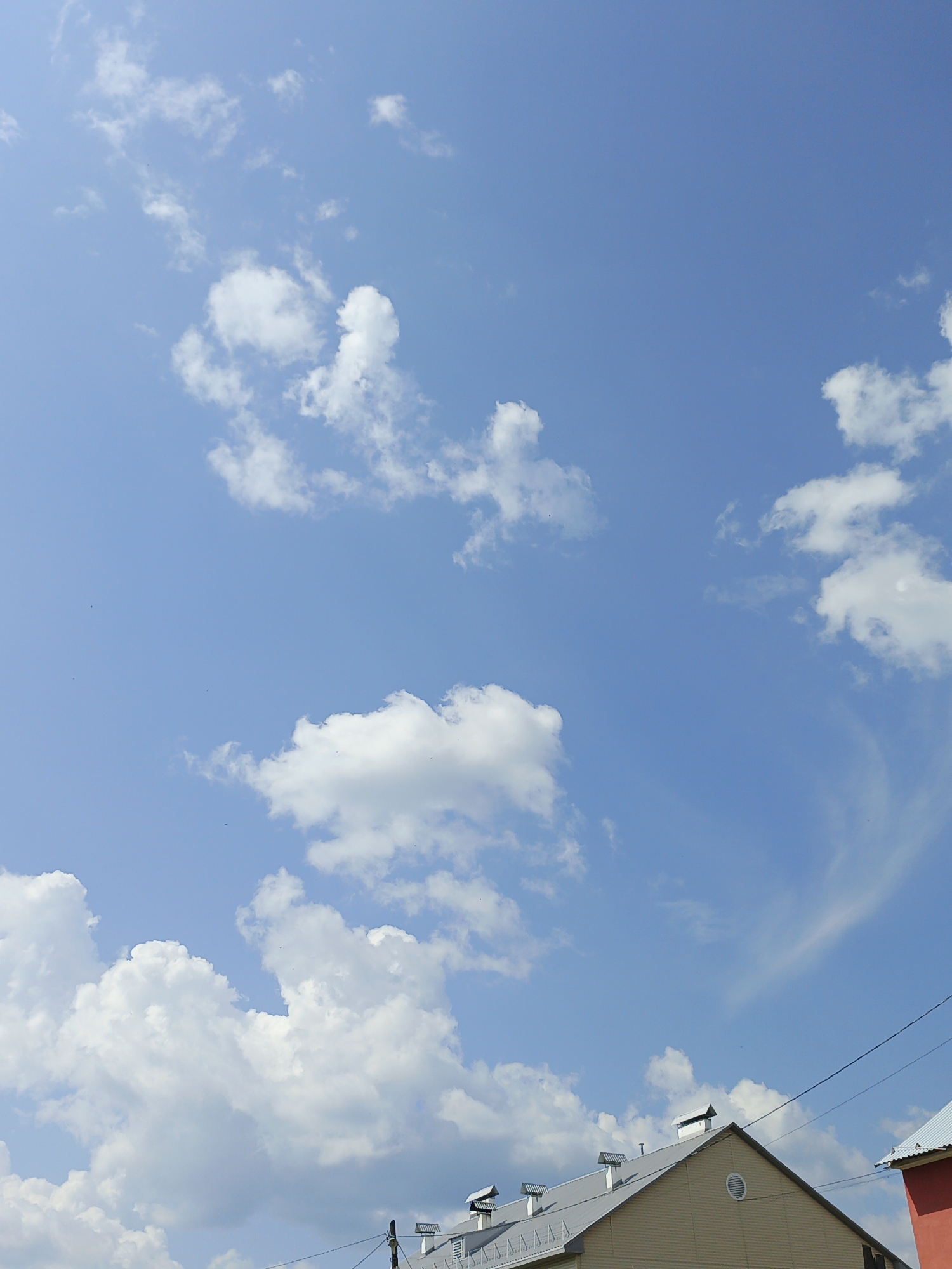 A bright, sunny sky with scattered white cumulus clouds dominates the upper portion of the image. The lower portion reveals the rooftops of buildings, suggesting an urban or suburban setting. The buildings appear to be of light-colored construction, with visible rooflines and some architectural details like vents and chimneys. Telephone wires are also visible, stretching across the frame. There are no people or animals present in the image. The scene is static and depicts a clear day. The image is a photograph taken outdoors in Medvedyovka, Russia.