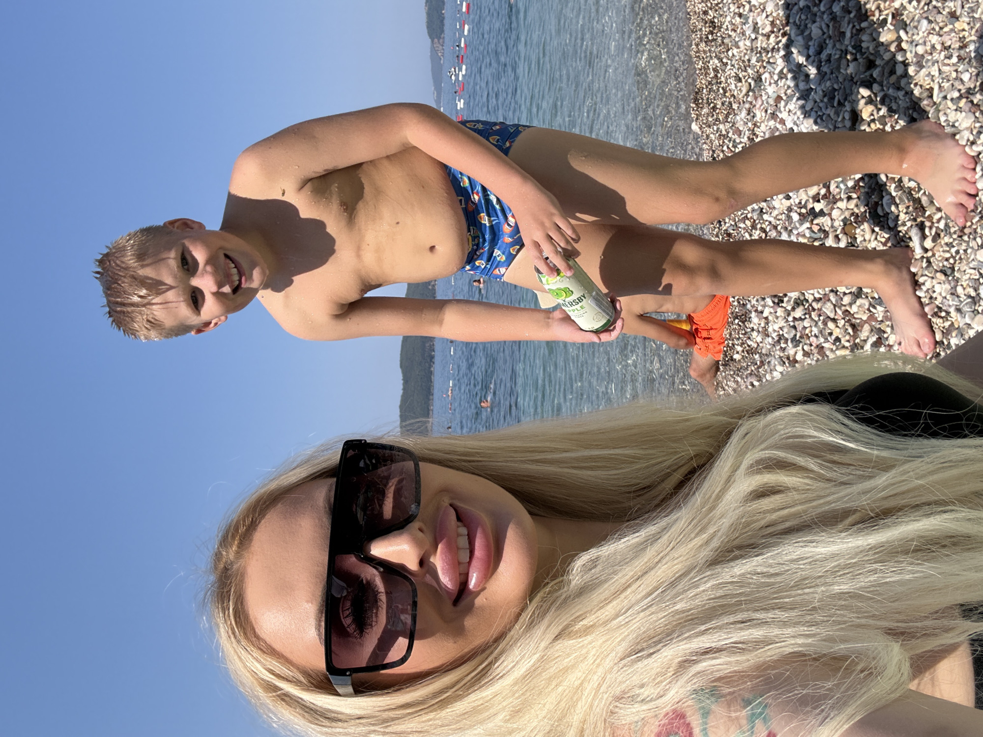 The media file is a selfie taken at a beach. In the foreground, a woman with long blonde hair and sunglasses is visible, her mouth slightly open, sticking her tongue out. She is wearing a black swimsuit. In the background, a young boy is also in the frame, partially submerged in the water. He is wearing blue swimming trunks and holding a can of what appears to be a beverage. The beach is covered in pebbles, and the ocean stretches out behind them, with boats and distant land visible. The sky is clear and blue, indicating a sunny day. The overall mood appears relaxed and joyful, suggesting a vacation or a day spent enjoying the outdoors. The can the boy is holding has visible text that reads "HERSHEY APPLE".
