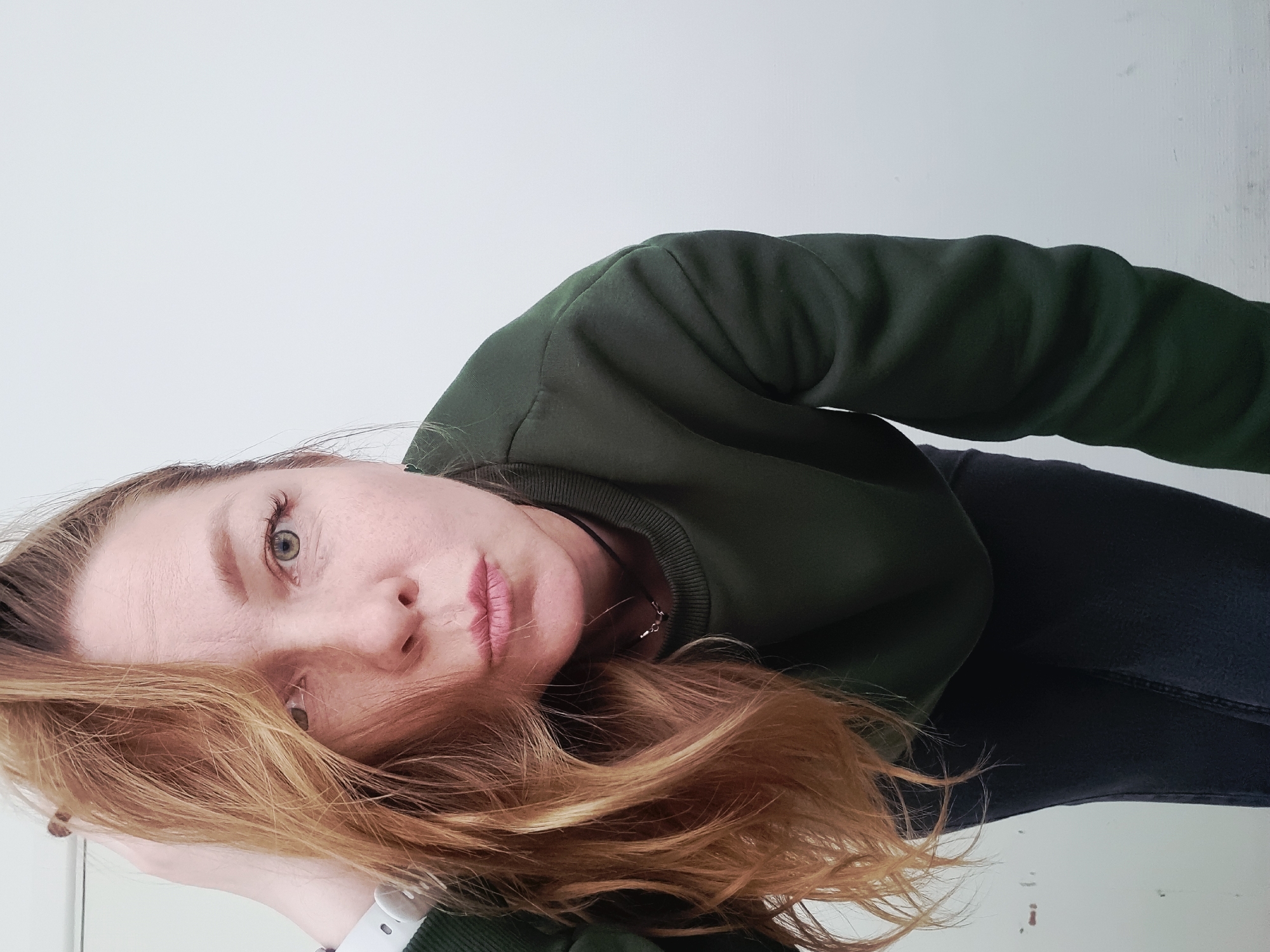 A woman is shown in a close-up, tilted portrait. She has fair skin, light-colored eyes, and light brown to blonde hair. She is wearing a dark green sweatshirt and a watch with a white band. The background appears to be a plain white wall. The location is not discernible as Kemul, Russia without additional context.