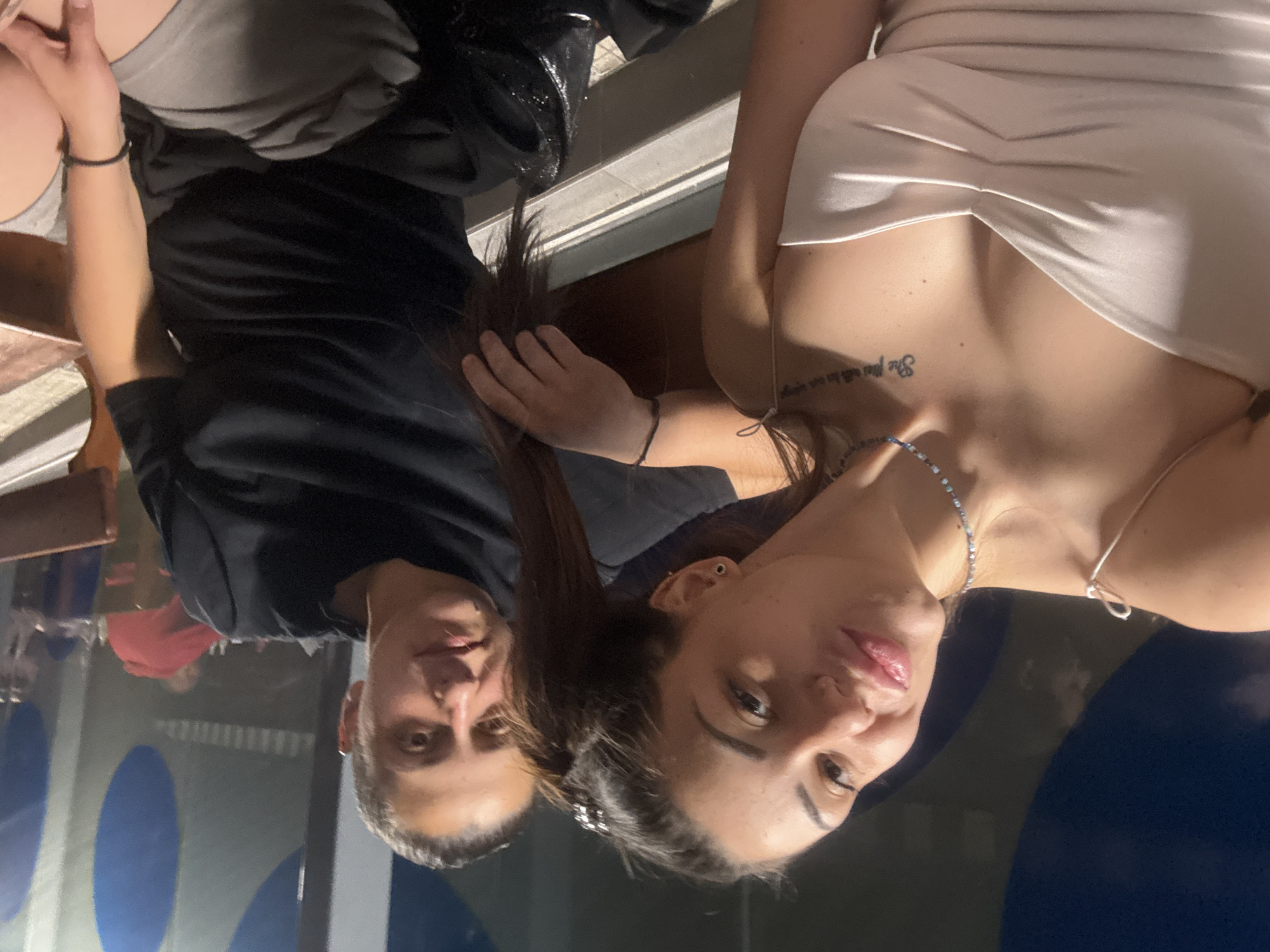 Two individuals are posing for a photograph in what appears to be an indoor setting. The individual on the left is wearing a black shirt and grey shorts. The individual on the right is wearing a beige dress and necklace, with a tattoo visible on her chest. The background features blue circles, suggesting a decorative element. The location might be in Sofia, Bulgaria, although no definitive landmarks confirm this.