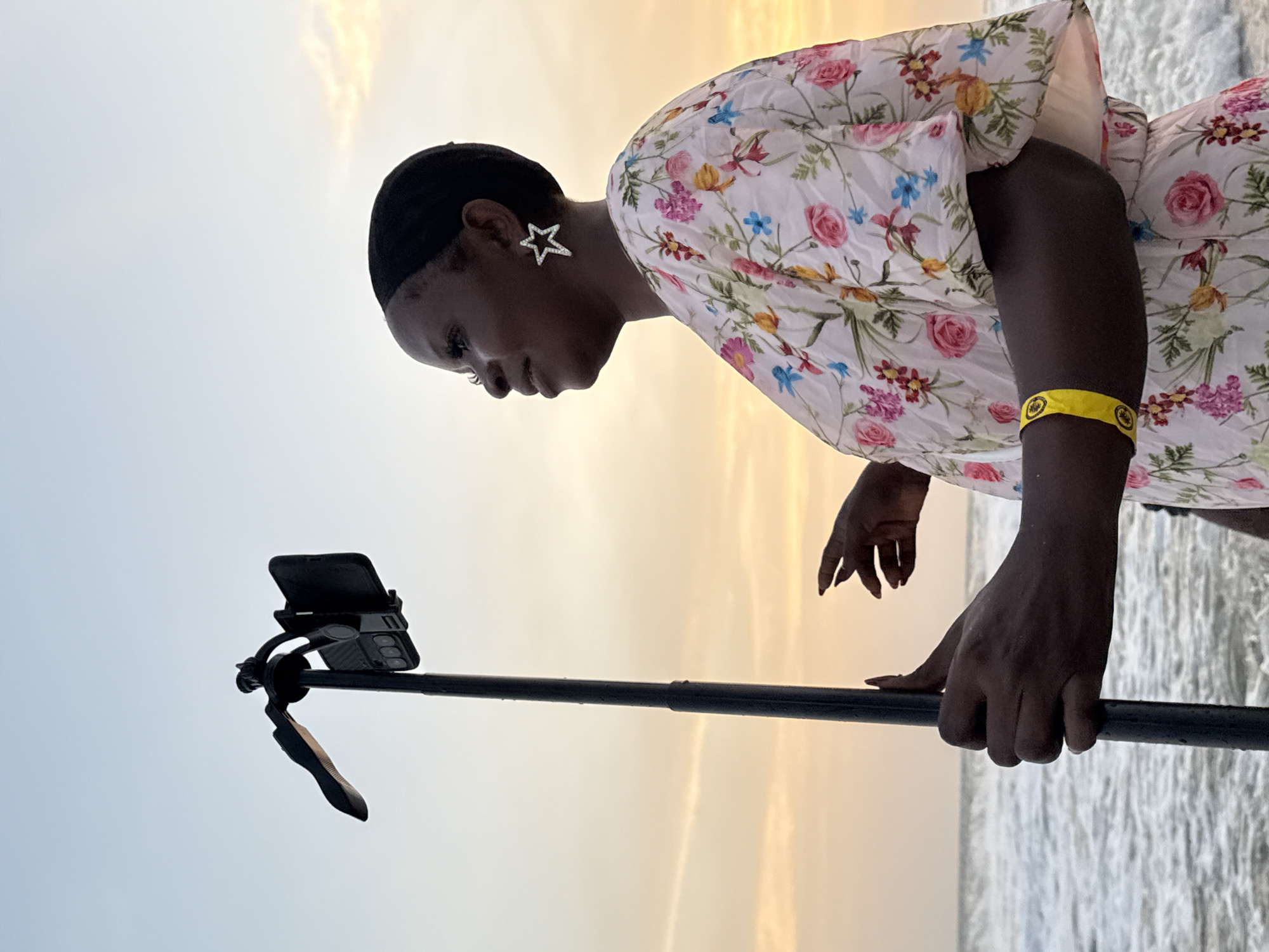 A low-angle, portrait-oriented shot shows a dark-skinned woman wearing a floral-patterned dress with short sleeves, a head covering, and star-shaped earrings. She is holding a selfie stick with a smartphone attached. The background shows a gradient of light blue to golden-yellow sky and a body of water, likely a beach or ocean, suggesting an outdoor location. The location is in Mopo Onijebu, Nigeria.