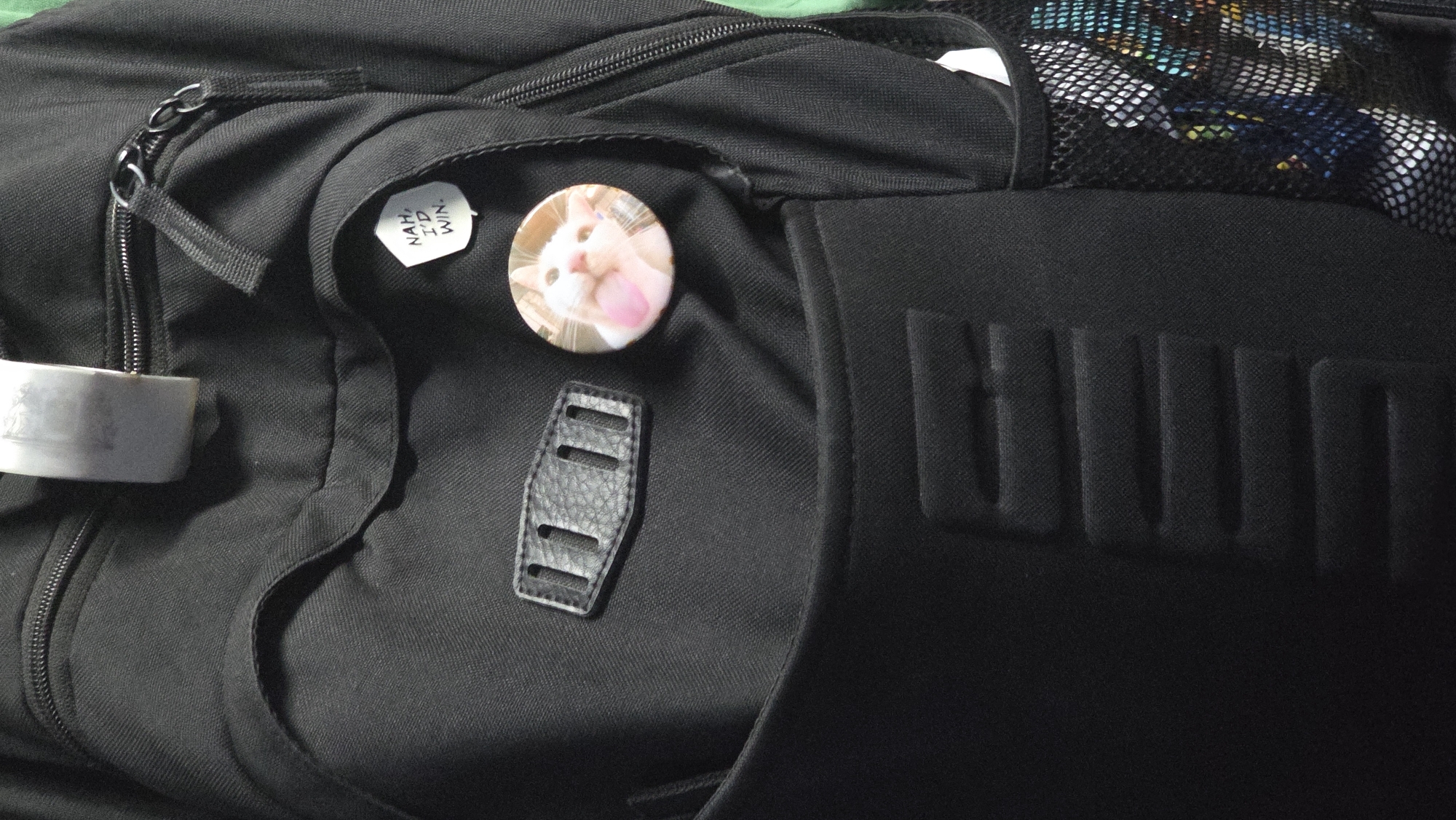A close-up, eye-level shot captures the front of a black backpack. The backpack is made of textured fabric and features several pockets and zippers. A prominent detail is a circular button pinned to the upper front pocket, which displays an image of a white cat with its tongue out. Above this, a smaller hexagonal button reads "NAH, I'D WIN." A leather-like tab with three vertical slits is also attached to the backpack. To the right, a black mesh side pocket reveals glimpses of colorful objects within. The scene is lit from above and to the right, casting subtle shadows. The image provides no information regarding location, including Riga, Latvia.