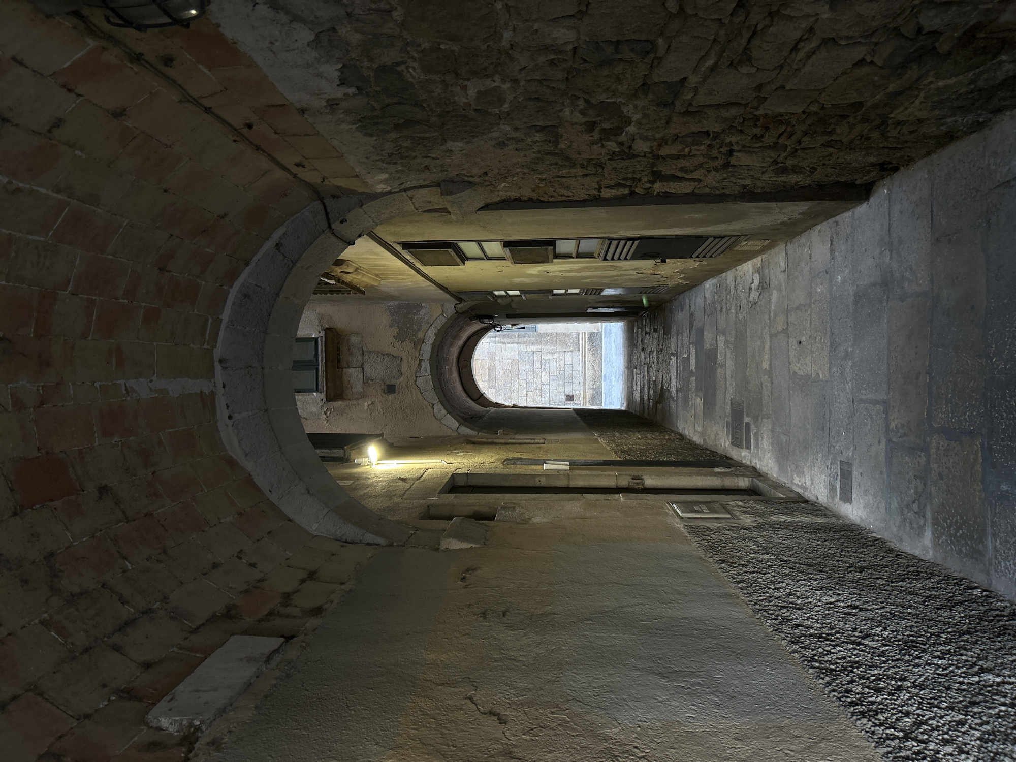 The image depicts a stone-paved alleyway in Girona, Spain, characterized by an arched ceiling constructed from reddish bricks and rough-hewn stones. The alley is narrow and curves slightly, with textured walls on either side. The left wall is predominantly rendered in a pale, rough stucco, with a band of dark, pebbled material running along its base. The right wall features exposed, irregular stonework. A single bright light fixture illuminates the left wall, casting a warm glow. Further down the alley, another archway is visible, leading to an area with more stone buildings and a lighter pavement. The scene appears quiet and devoid of people, suggesting it might be early morning or late evening.