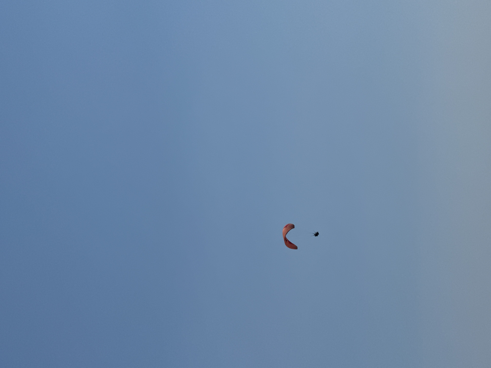 A lone paraglider, clad in dark clothing, soars against a clear, pale blue sky. The paraglider's wing is a vibrant red, catching the light and creating a stark contrast with the vast expanse of the sky. The paraglider appears to be high in the air, with no discernible land features or cityscapes visible, suggesting an open area likely for aerial sports. The image does not provide specific geographical markers that would pinpoint the location to Boreti, Montenegro, or any other urban environment, as the focus is exclusively on the paraglider and the sky.