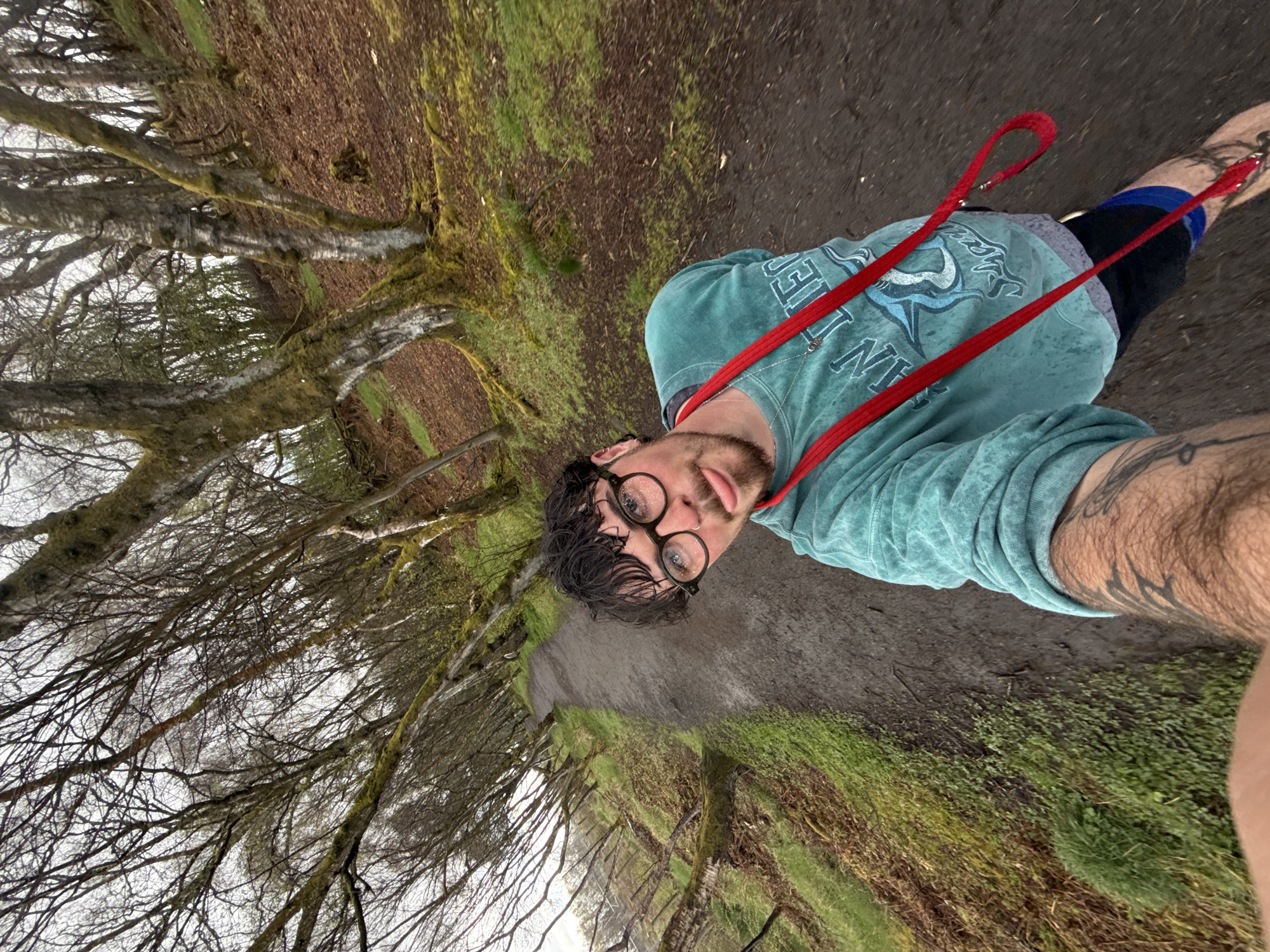 A selfie photograph shows a man on a trail in an unknown location, city, and state. The man is wearing a teal shirt, black shorts, and round glasses. He is holding a red dog leash. The trail is dirt and surrounded by trees and greenery. The photograph is taken at an angle.