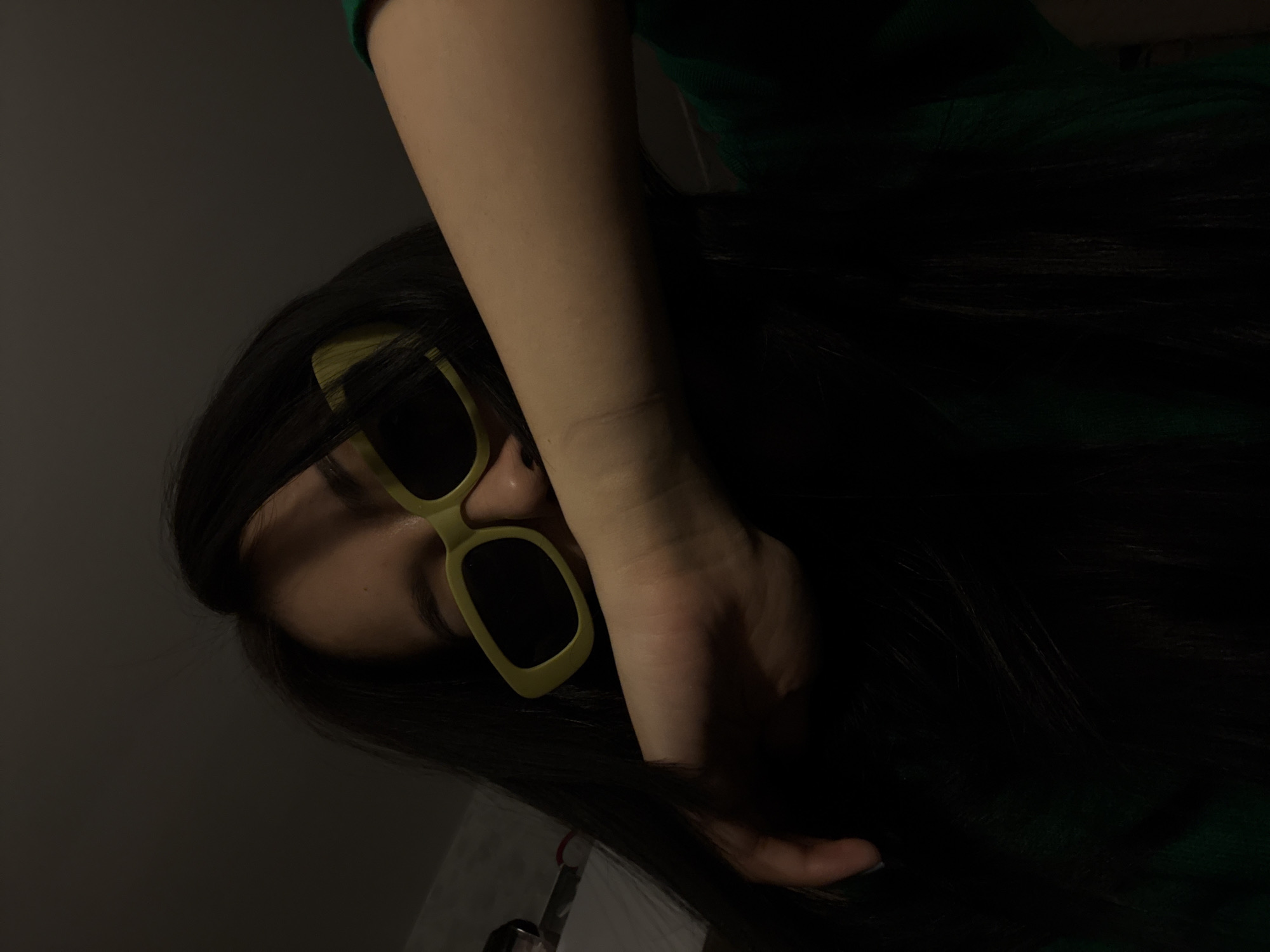 A person with dark hair and skin wearing green sunglasses and a green shirt covers part of their face with their hand. The scene is poorly lit. The image does not contain sufficient information to identify the location in Votkinsk, Russia.