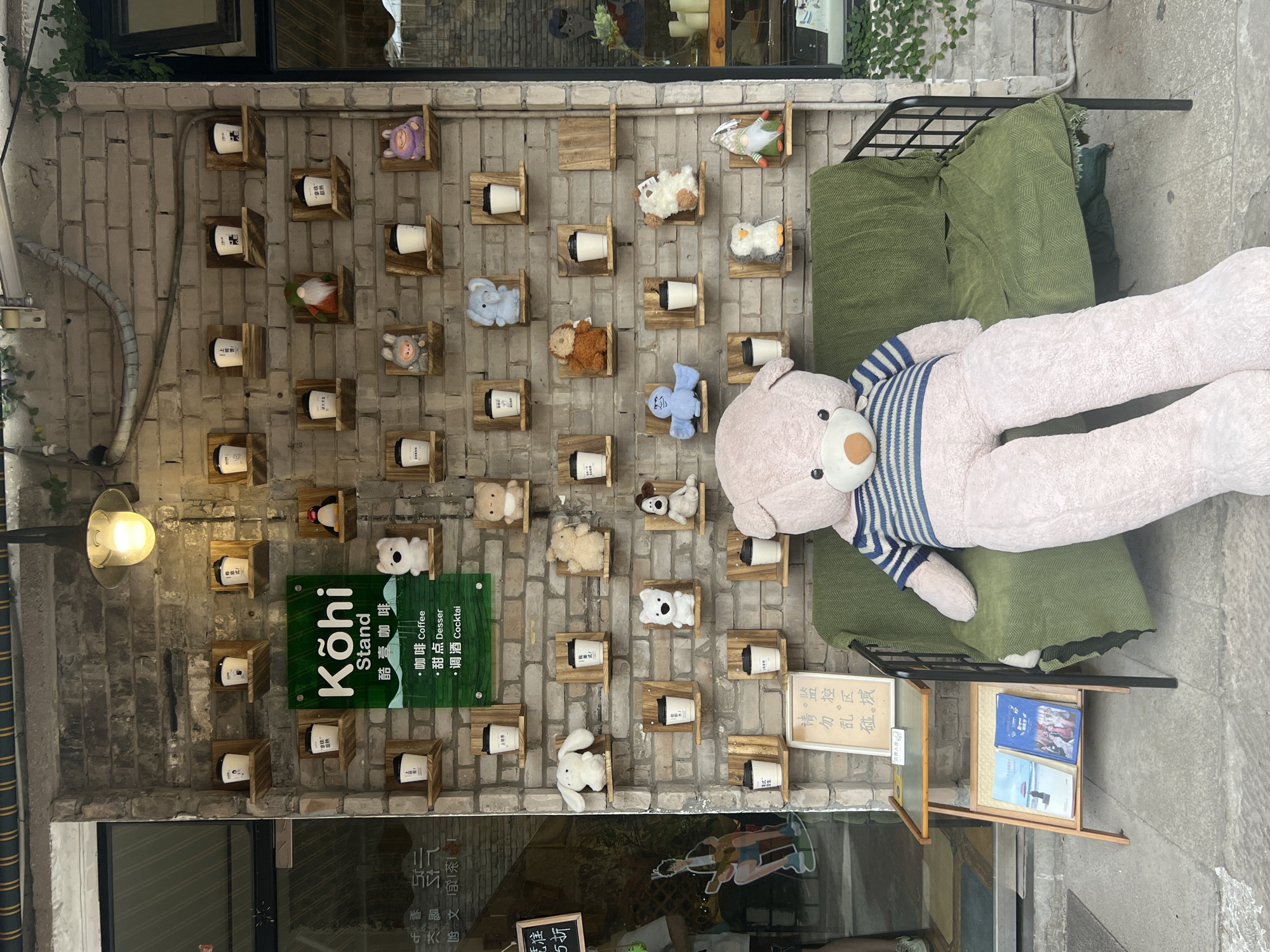 This is a medium shot of the exterior of a cafe in Yuzhong District, China, featuring a large plush teddy bear sitting on a green outdoor sofa. The teddy bear is light pink and wearing a blue and white striped sweater. It is positioned to the right of the frame, facing slightly to the left. The sofa is covered with a green, chevron-patterned throw.
Behind the sofa, a brick wall serves as a backdrop for a display of small shelves holding coffee cups and plush toys. A prominent green sign with "Kōhi Stand" in white lettering is visible, along with Chinese characters and text indicating "Coffee," "Dessert," and "Cocktail." Several wooden shelves are arranged in a grid on the brick wall, each displaying a white coffee cup with a black lid, and some also feature small plush toys.
To the left of the teddy bear and sofa is a small wooden side table with two books displayed on it. A framed sign with Chinese characters is placed on top of the side table. Further to the left, a large glass window with reflections is visible, and a portion of a doorway can be seen. Green vines are growing along the upper part of the brick wall. The lighting suggests it is daytime.