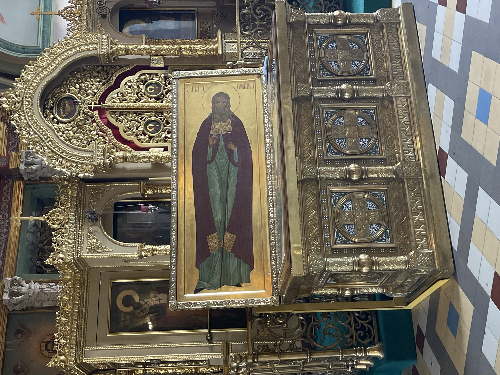 The image shows an ornate, gilded altar within the Trinity Lavra of St. Sergius in Sergiyev Posad, Russia. A large icon depicting a bearded saint in religious vestments is displayed on a gilded reliquary. The reliquary itself is intricately carved with religious motifs and sits on a tiled floor. The background features gilded architectural elements and religious paintings, typical of Orthodox Christian churches.