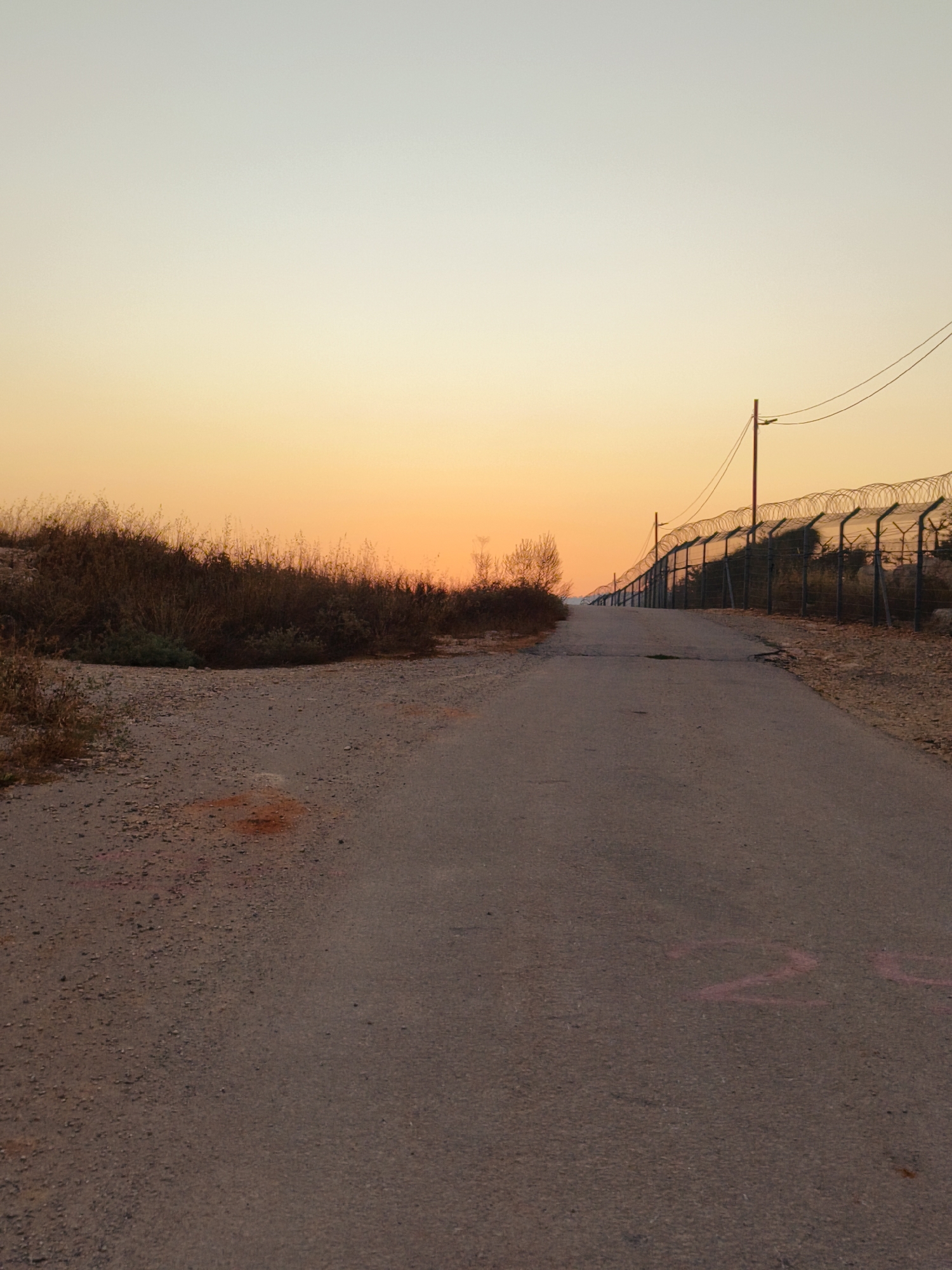 The image depicts a paved road leading towards a barbed wire fence in the Lachish Regional Council, Israel, at sunset. The sky is a gradient of soft orange and pale yellow. To the left of the road, dry brush and sparse vegetation line the embankment. The barbed wire fence, supported by metal poles, stretches along the right side of the road, appearing to demarcate a border. Electrical wires are visible above the fence. The road itself is asphalt and shows some wear and tear, with pink markings on its surface. The overall scene suggests a border or restricted area under a twilight sky.