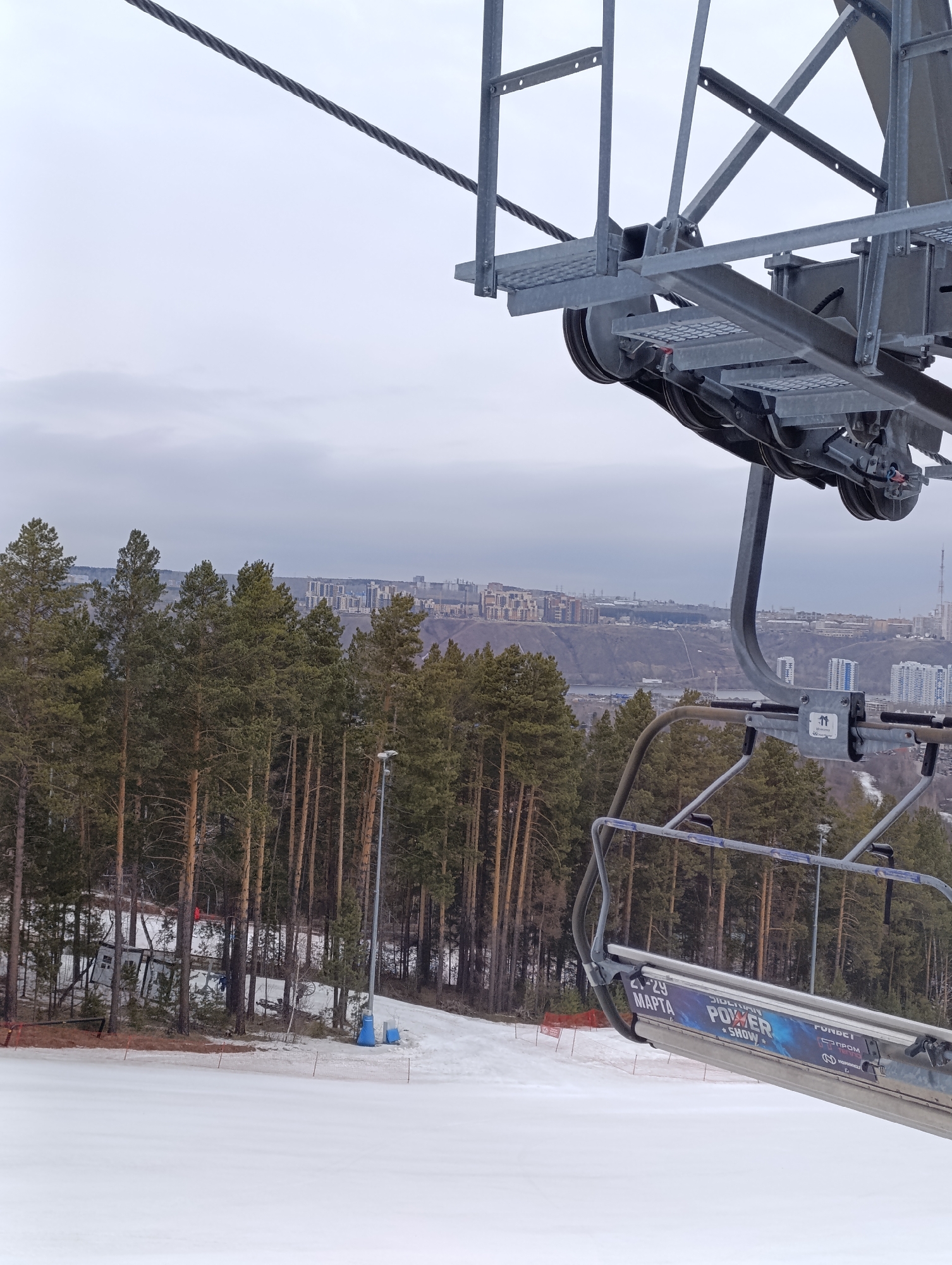 The image presents a view from a ski lift chair overlooking a snow-covered slope in the city of Krasnoyarsk, Russia. Tall evergreen trees line the edge of the ski run, and in the distance, urban buildings are visible against a cloudy sky. The ski lift chair itself is in the foreground, with its metal frame and advertisement for "Siberian Power Show 21-29 MAPTA". The cable of the ski lift is also prominent in the upper part of the frame. The snowy slope shows tracks and some fencing, suggesting it is an active ski area.