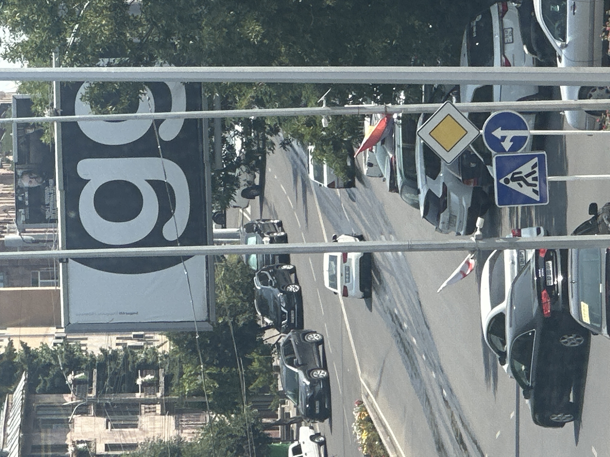 The image captures a street scene in Gyumri, Armenia, with a focus on a billboard featuring the letters "gg" in large white font against a black background. Several cars are visible on the road, including SUVs and sedans, some of which are in motion. Traffic signs, including a pedestrian crossing sign, a diamond-shaped yellow sign, and a directional arrow sign, are present. Buildings and trees form the background, with the architecture suggesting an urban environment. The overall impression is a snapshot of everyday life in a city.