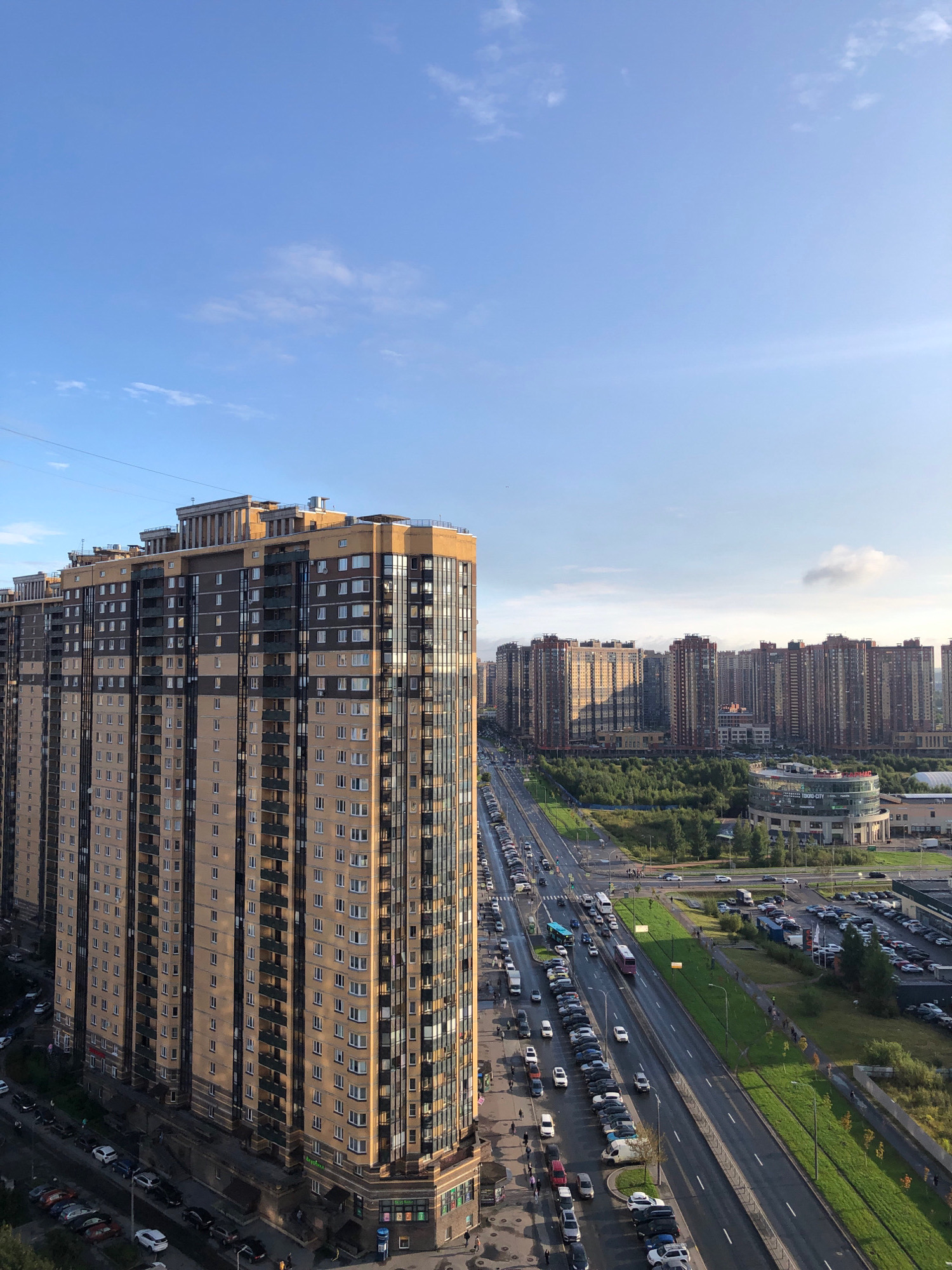 An aerial shot captures a dense urban landscape under a partly cloudy sky, with a focus on a tall, multi-story residential building on the left. The building features a beige and dark grey facade with numerous windows and balconies, extending vertically across the frame. To the right of this building, a wide, multi-lane road runs diagonally into the distance, lined with parked cars on both sides. A row of trees separates the road from a large, circular commercial building with glass-paneled sections, identifiable by the signage "TORDO-CITY". Further into the background, a dense cluster of similar tall residential buildings stretches across the horizon, interspersed with green spaces. The scene suggests a modern, developing urban environment in a Russian city.