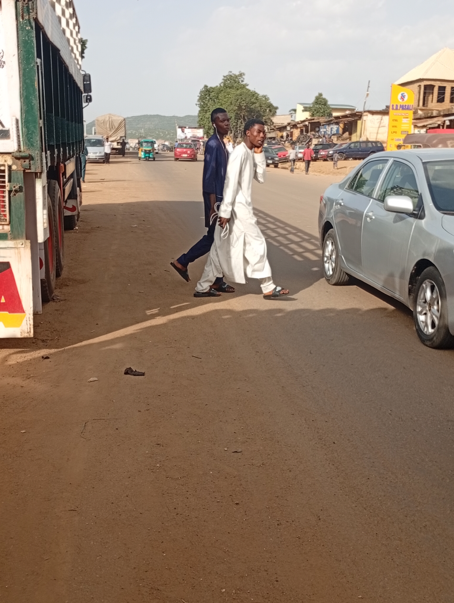 The image depicts a dusty road scene in Rafin Par, Nigeria. In the foreground, two young men are walking across the road. The man on the left is wearing a dark blue garment and a dark cap. The man on the right is wearing a white traditional outfit. To the left of the frame, a large white truck with a checkered pattern on its side is parked. In the background, several vehicles, including cars and a small turquoise auto-rickshaw, are visible on the road. Buildings with corrugated iron roofs line the side of the road. The environment appears dry and sunny with a clear sky.
