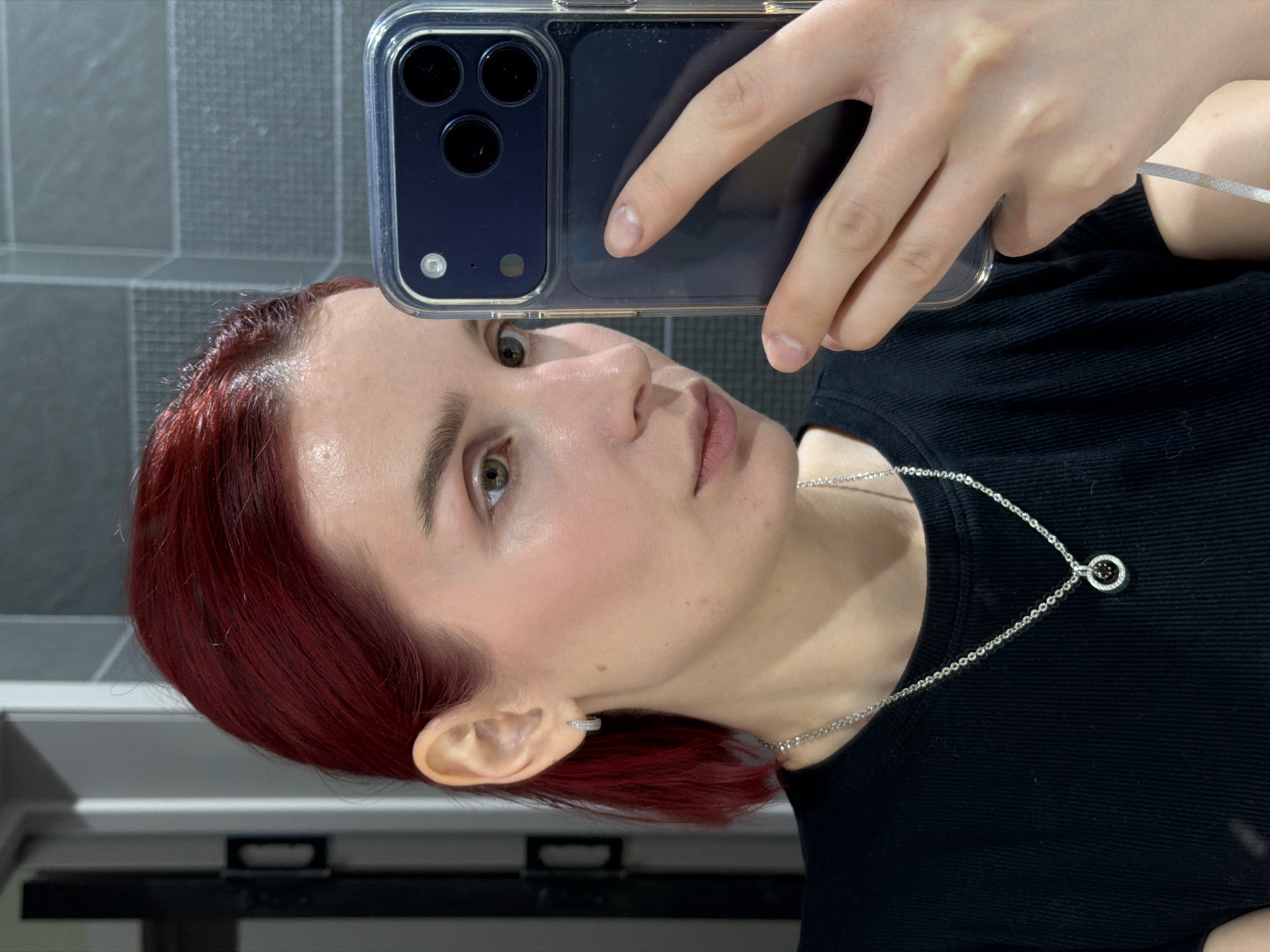 The media file is a rotated mirror selfie taken indoors, featuring an individual with red/auburn hair, fair skin, and light-colored eyes (green/hazel). The person is wearing a black ribbed t-shirt or top and a silver chain necklace with a circular pendant, which has a small dark or reddish stone in its center. A small silver hoop earring adorns their right ear.
The individual is holding a dark blue smartphone, enclosed in a clear case, with their right hand to capture the reflection. The phone's multi-lens camera array and flash are clearly visible. The person's gaze is directed towards the phone's camera in the reflection, with a neutral and calm expression. Faint red marks are visible on the forehead.
The background reflected in the mirror consists of a dark grey or black tiled wall with a grid pattern, suggesting an indoor setting such as a bathroom or dressing area in Chaikovsky, Russia. Below the tiled wall, some dark, horizontal structures, possibly a counter or shelf, are discernible. The lighting is bright and evenly distributed. No visible text is present in the image.