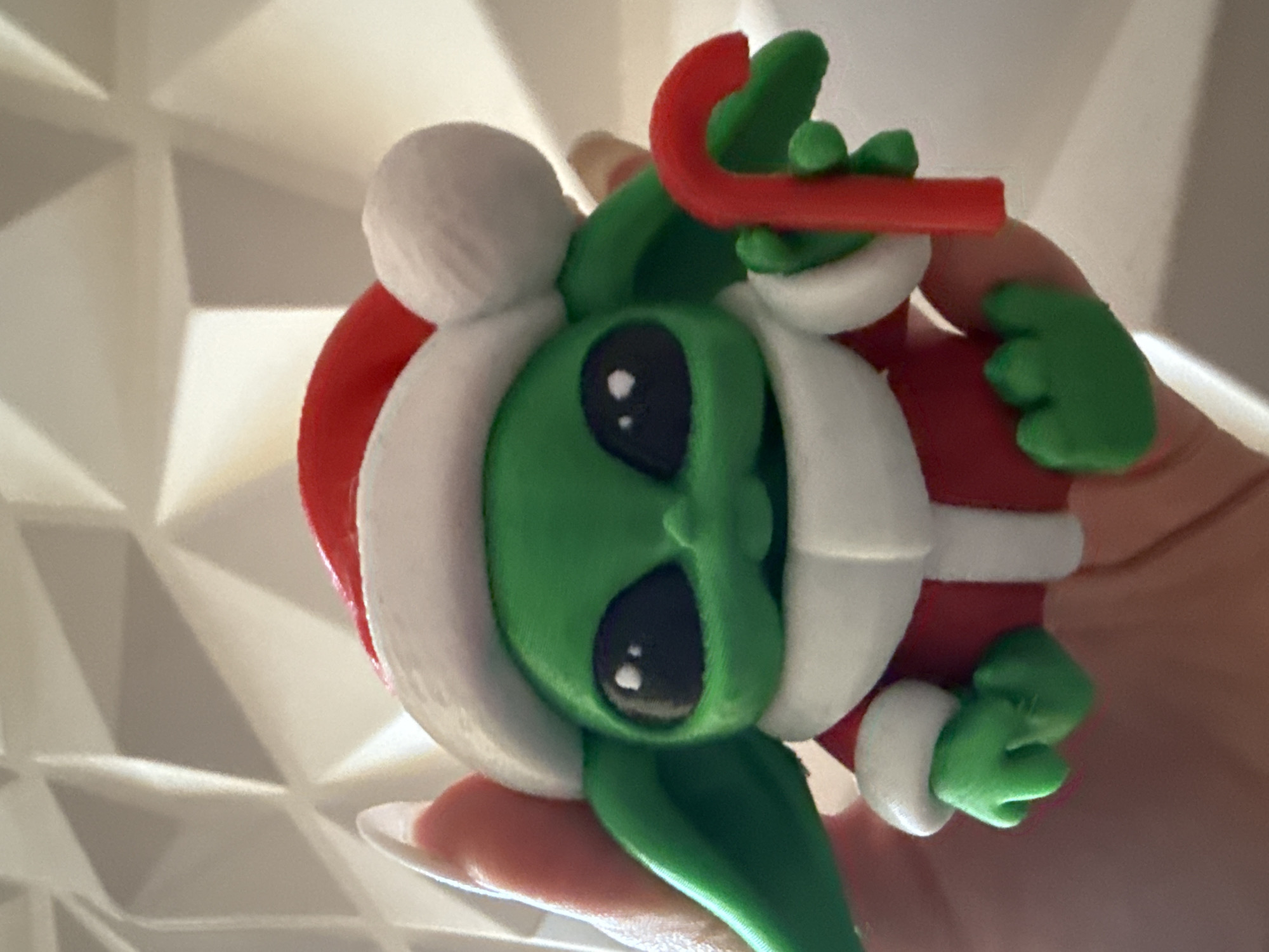 A hand holds a handmade, green-skinned alien in a red Santa suit with white trim and a white pompom on the hat. The alien has large, black eyes and is holding a red candy cane with green leaves. The background is a textured white surface.