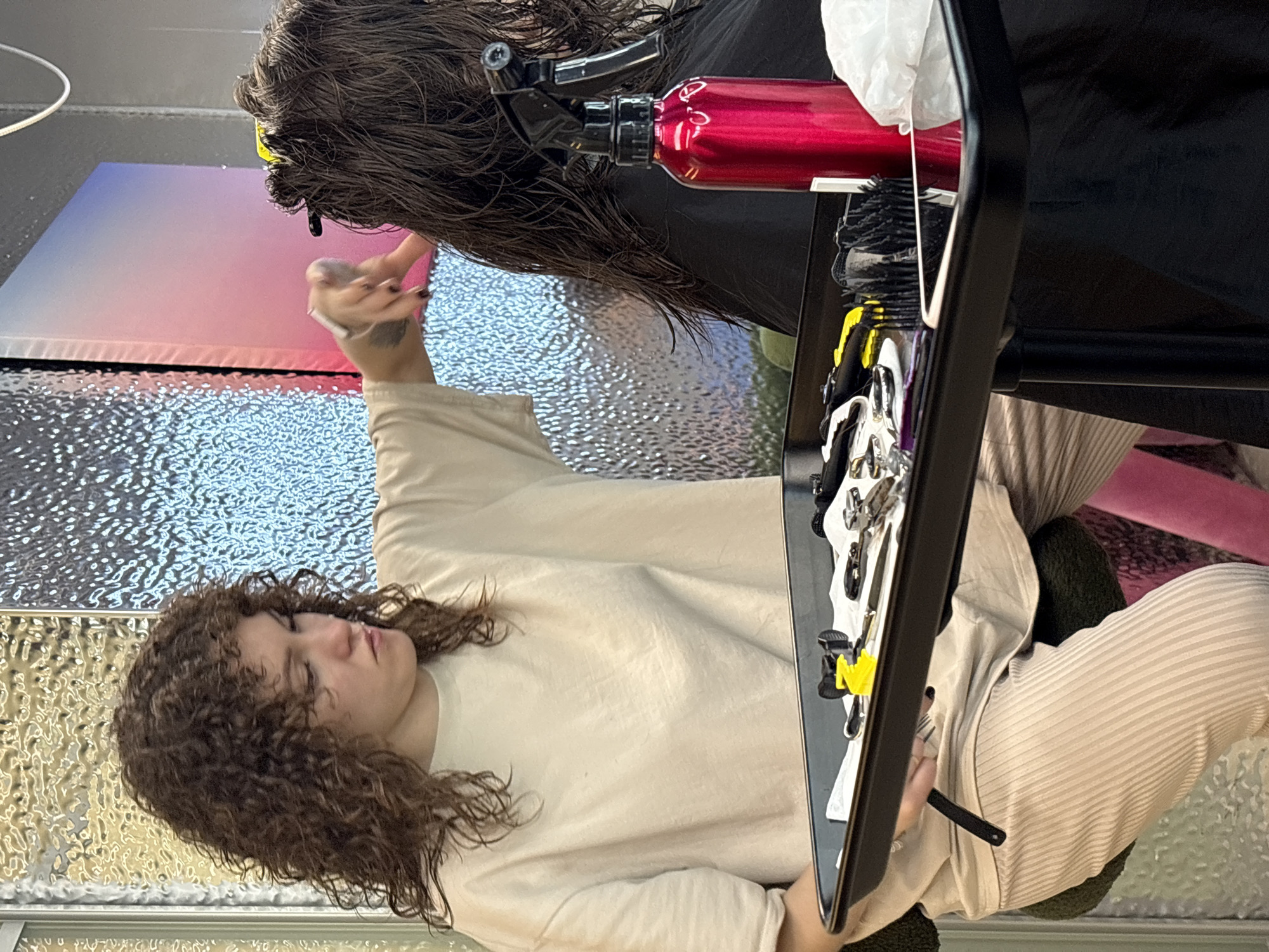 A woman with curly brown hair, wearing a light-colored t-shirt and striped pants, is seated in a salon and appears to be cutting the hair of another person, whose back is to the camera. She is holding a pair of scissors in her right hand and is reaching towards the other person's wet hair. A black tray in front of her contains various hair styling tools, including scissors, combs, and a red spray bottle. The background features a textured wall with a gradient of pink and blue colors. The scene appears to be a hair salon.
