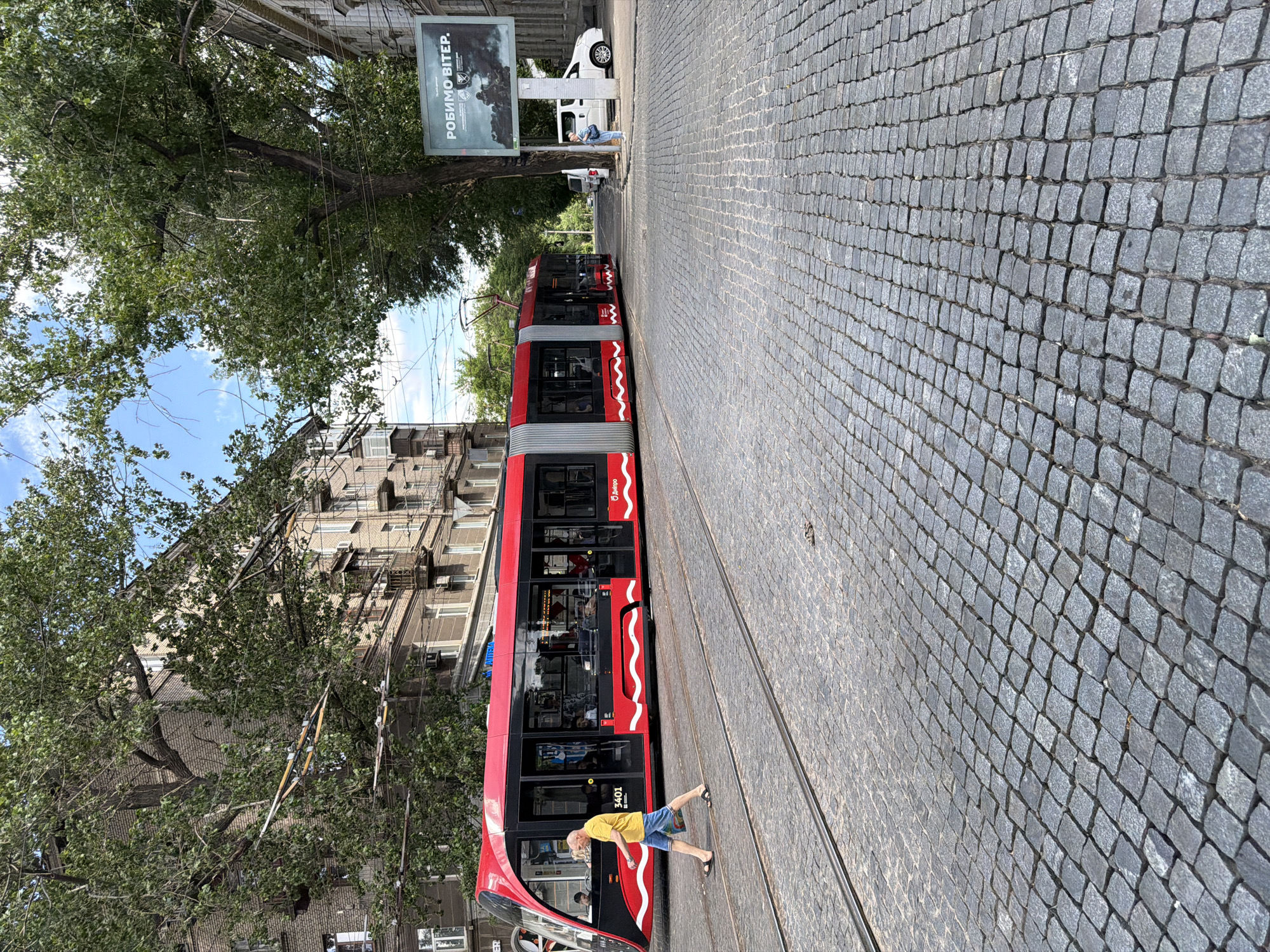 In Dnipro, Ukraine, a modern red and black tram with the number 3401 is traveling down a cobblestone street. A man in a yellow t-shirt and blue shorts is walking across the tram tracks, carrying a bag. The street is lined with old brick buildings and large trees. A billboard with Ukrainian text reading "РОБИМО ВІТЕР" (We Make Wind) is visible in the background, along with a white van and another vehicle. The sky is partly cloudy.