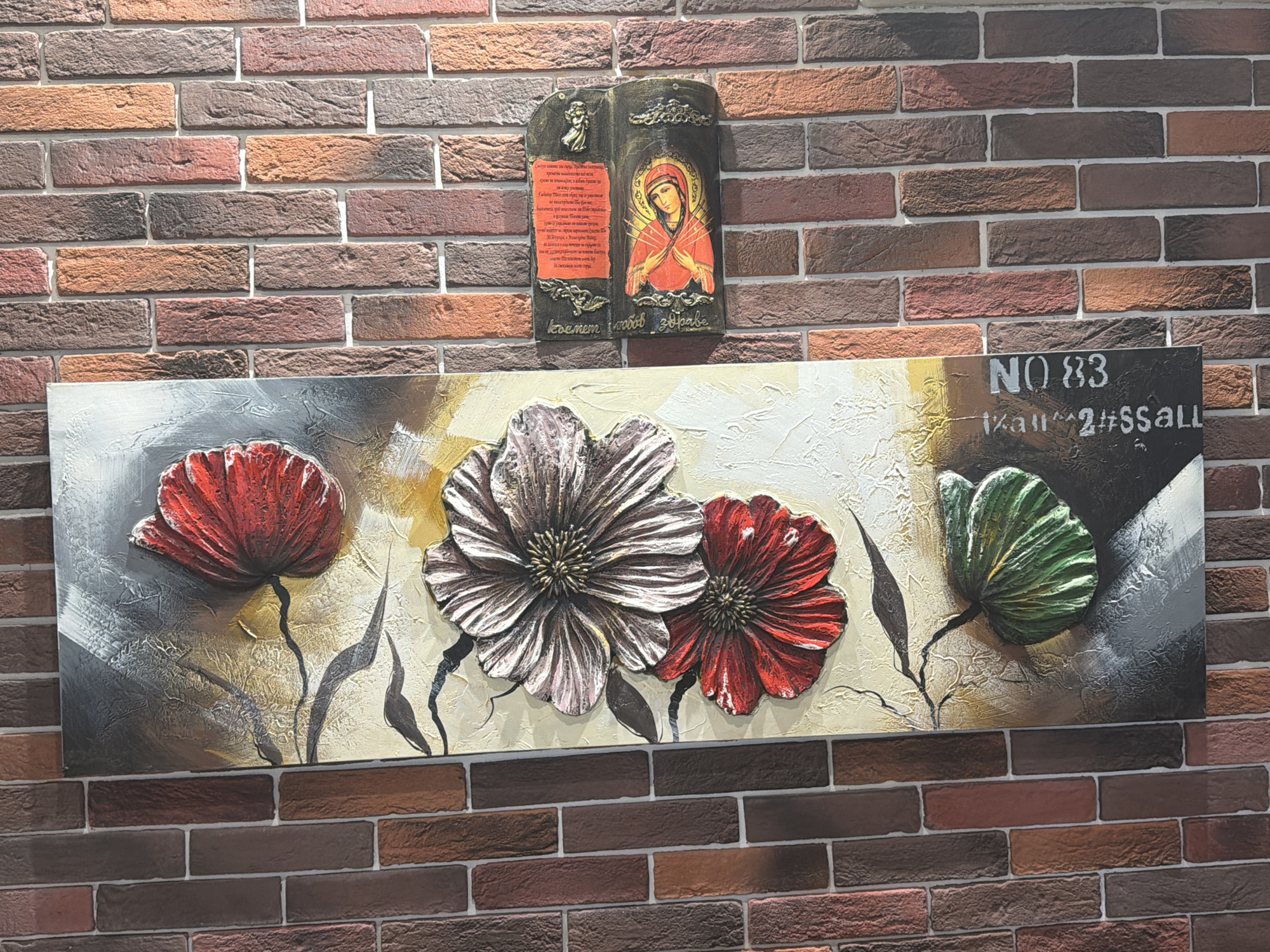 A wall in Plovdiv, Bulgaria, is decorated with three items. The wall is made of reddish-brown bricks with a white grout. The top item is a framed religious icon featuring a Madonna-like figure in red with some text in a non-Latin alphabet. Below the icon is a larger framed piece of art with three flowers and a green leaf. The flowers are red, white, and red, and the art appears to be a three-dimensional painting. There is some graffiti-like writing at the top right of the large art, with the number "83" followed by more text in a non-Latin alphabet.