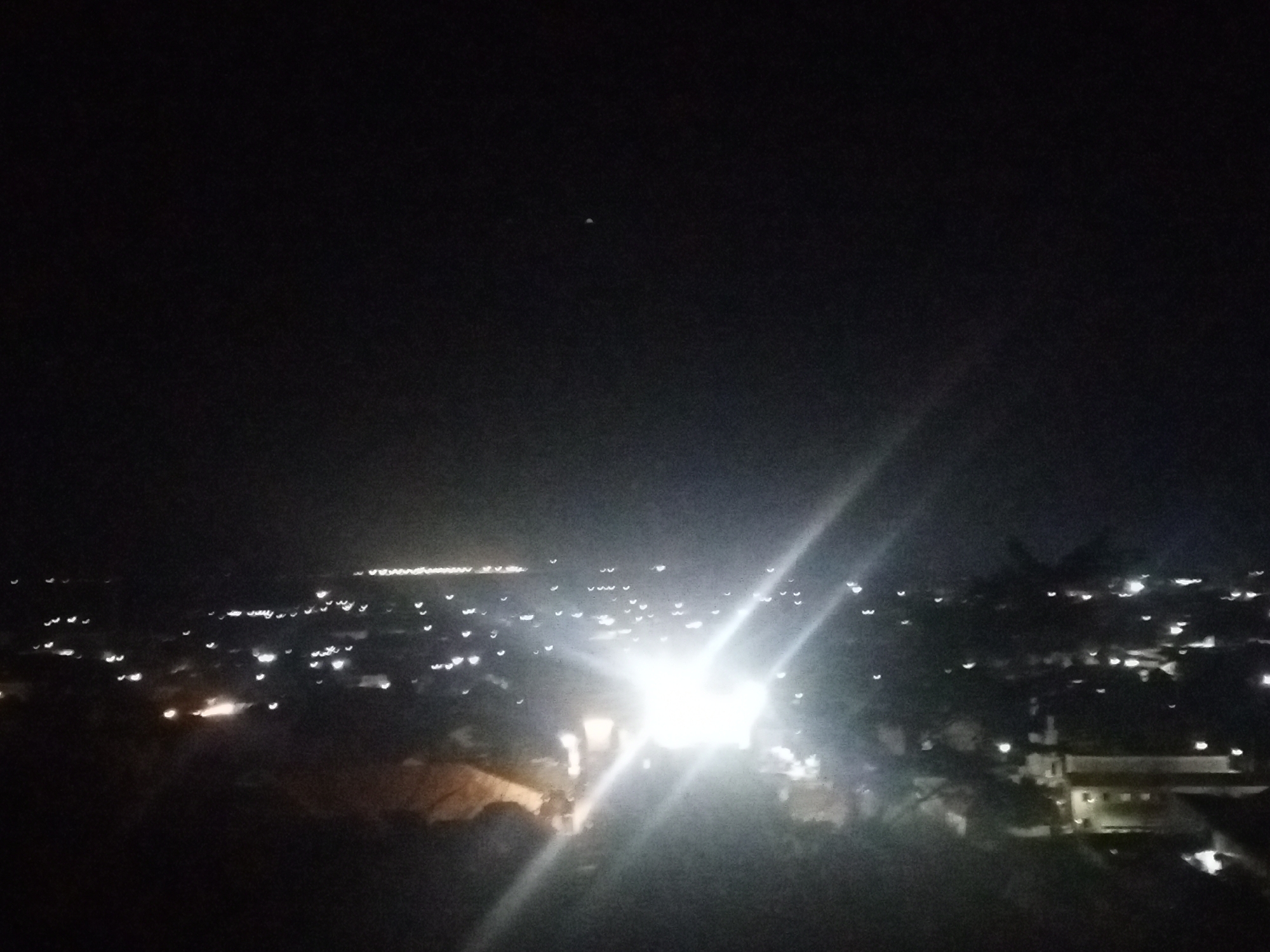 The image shows an elevated night view of a city, most likely taken from a high point or building. The city is identifiable by the scattered lights of buildings and street illumination. There is a particularly bright light source closer to the foreground. The sky above is dark, with a small moon or other celestial body visible in the upper part of the frame. Some trees and foliage are visible in the foreground. The image does not provide any specific information about the location of the city in relation to the city of Unknown, Unknown. There are no people visible in this image.