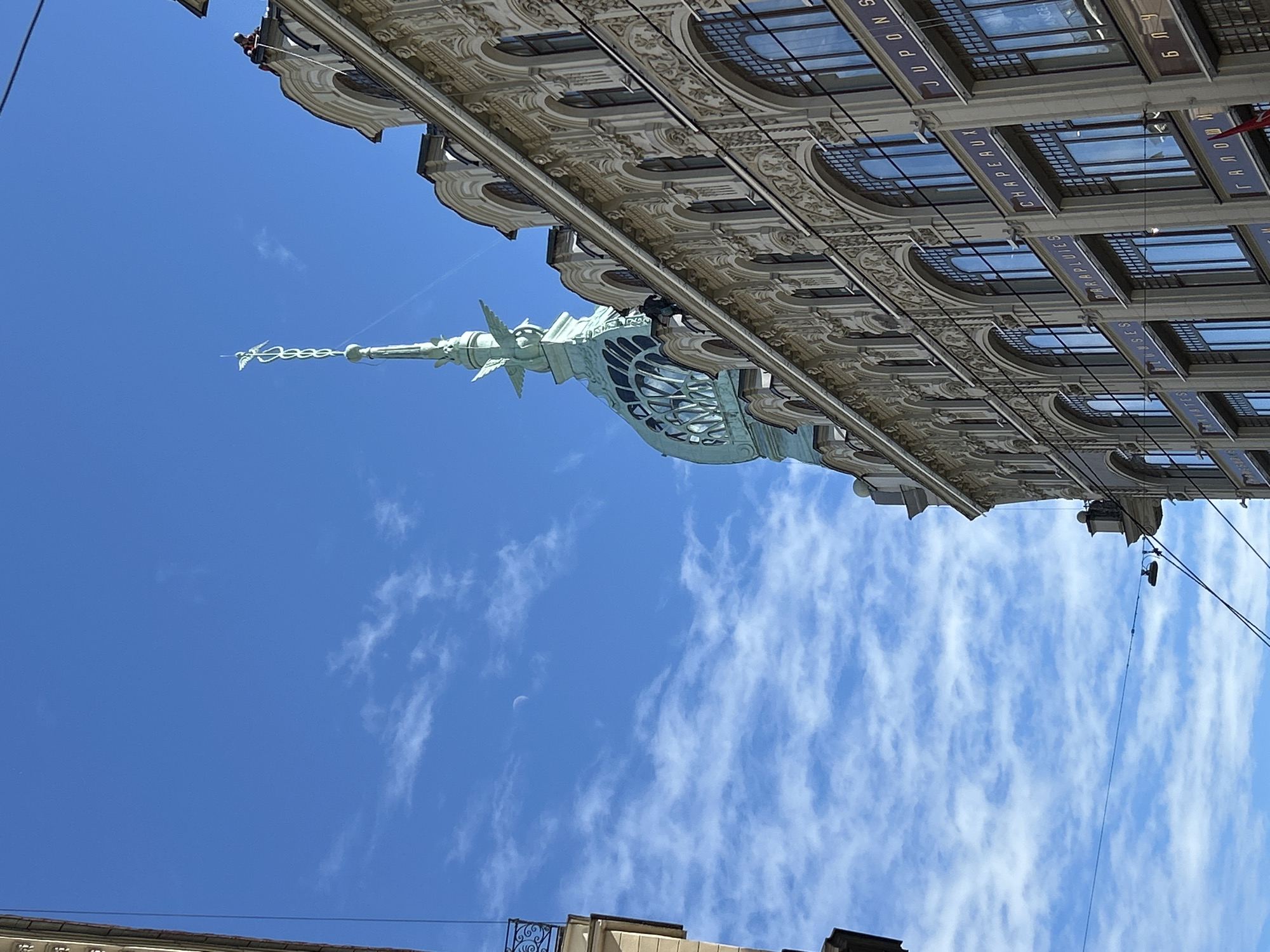 A low-angle, close-up, exterior shot captures the ornate facade of a historic building against a bright blue sky dotted with wispy clouds. The building features a cream-colored, intricately decorated exterior with multiple arched windows. Above, a prominent turquoise spire, topped with a delicate, metallic figure, rises dramatically. A person in an orange jacket is visible near the top of the building on the roof, seemingly engaged in work. The scene is framed by the edge of another building on the left and overhead wires, suggesting an urban environment.