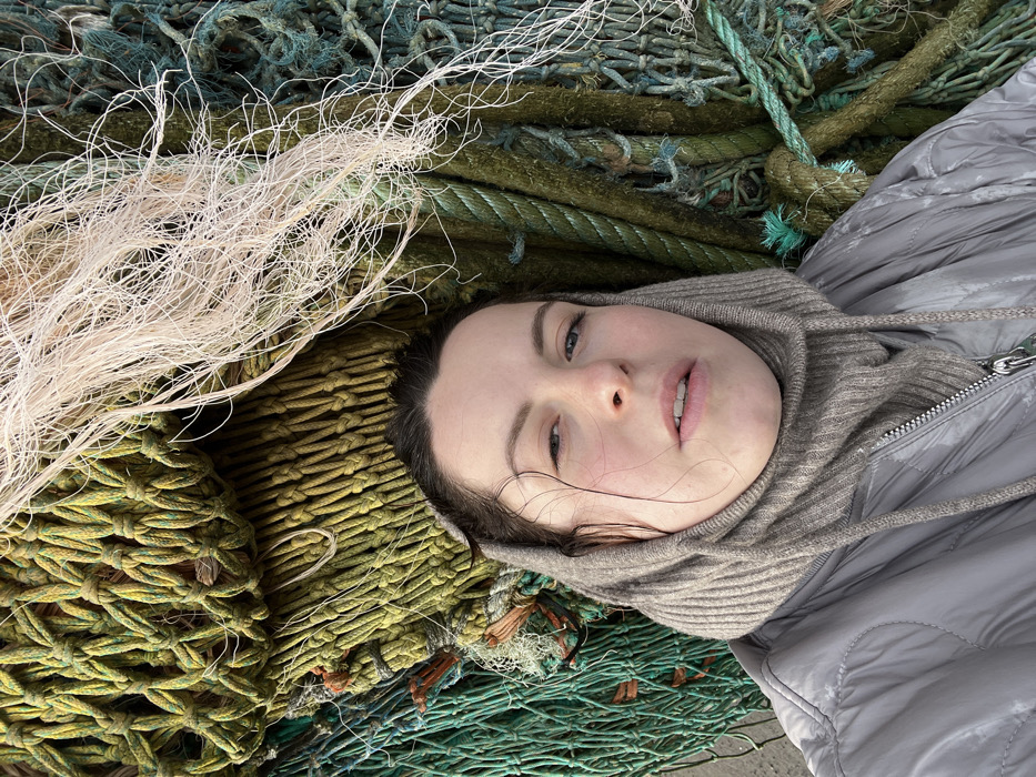 A female individual is captured in a close-up portrait, situated in Charmouth, United Kingdom, against a dense, textured backdrop of discarded or stored fishing nets and ropes. The subject, appearing to be an adult female, is seen from the chest up, looking directly at the camera with a neutral expression, eyes open, and mouth slightly ajar. Her dark hair is somewhat damp or disheveled, with strands falling onto her forehead and temples. She wears a light grey, possibly padded or quilted, jacket with a zipper closure, and a ribbed, light brown or beige balaclava-style garment covering her head and neck. The background consists predominantly of tangled, weathered fishing nets and thick ropes in various shades of green, olive, and teal, indicating marine use and exposure to elements. A section of lighter, almost white, frayed fibrous material is visible at the upper right. The materials in the background are tightly packed and show visible knots, weaves, and signs of wear. The overall scene suggests a coastal or harbor environment where such marine equipment is present.