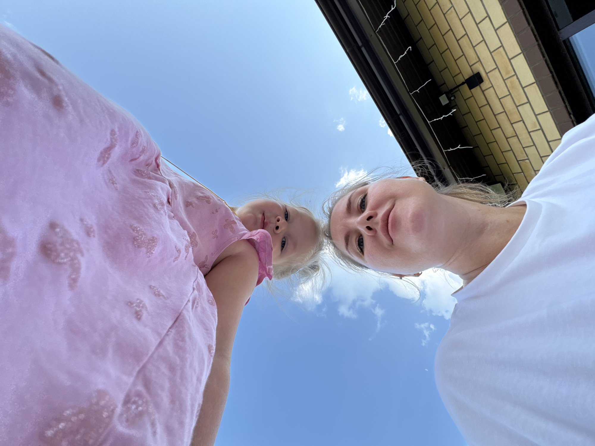From a low-angle perspective, the image captures a woman and a young girl against a backdrop of a clear blue sky. The woman, wearing a white t-shirt, is positioned to the right, her face close to the camera. Adjacent to her is a young girl in a pink dress. The backdrop includes a section of a building constructed of light-colored bricks. It's Ферма, Russia.
