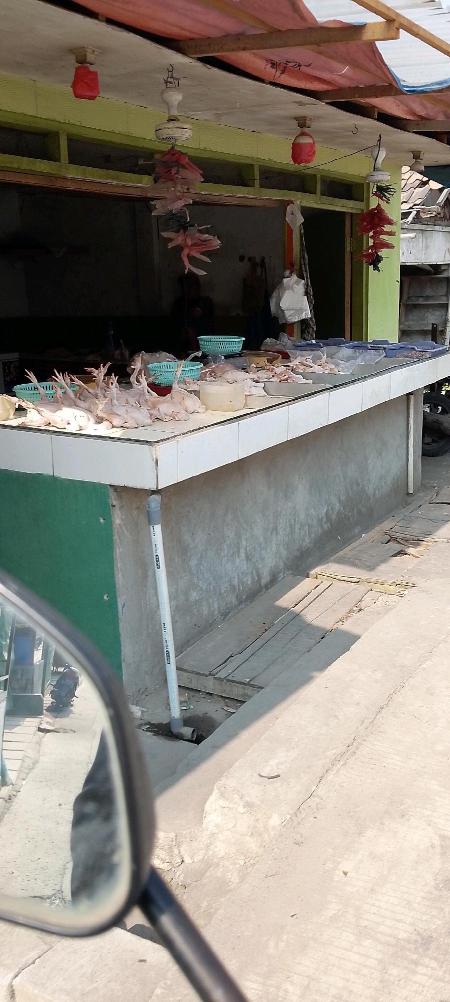 The image depicts an outdoor food stall, likely a butcher shop, in Babelan Kota, Indonesia. The stall features a long, tiled counter displaying numerous whole raw chickens, some arranged in small teal-colored baskets. In the background, the stall's interior is visible, with a person obscured by shadow. Various decorations, including red and pink paper lanterns and what appear to be hanging plastic strips, are strung from the ceiling. A PVC pipe runs vertically along the green-painted wall next to the counter, connecting to an outlet on the ground. The ground in front of the stall is paved, with some wooden planks laid out, and a motorcycle is partially visible in the foreground reflected in a mirror. The environment suggests a sunny day.