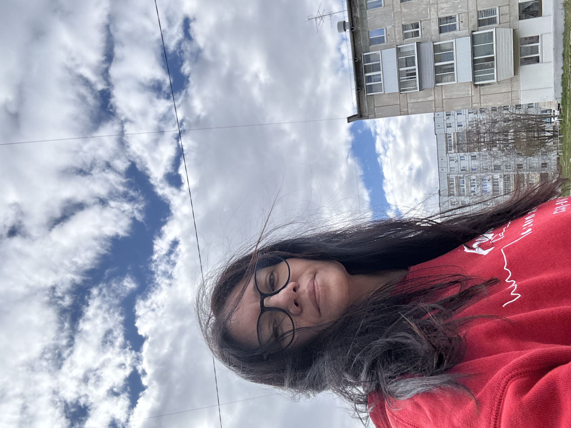 A selfie of a woman with long, dark hair and glasses, wearing a red hoodie, is taken outdoors in Chaikovsky, Russia. Behind her is a partially visible apartment building, typical of post-Soviet architecture, with multiple windows and balconies. The sky is visible with scattered clouds and patches of blue. Overhead power lines are also visible.