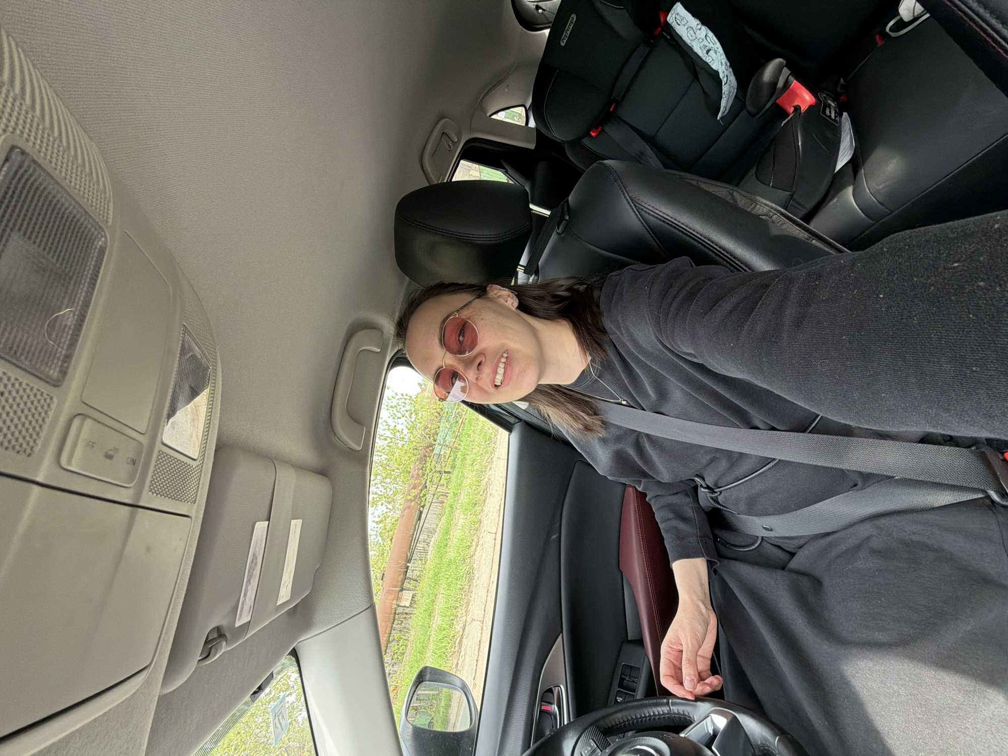 A woman wearing rose-tinted round glasses and a black dress is seated in the driver's seat of a car, wearing a seatbelt. The car has black leather seats with red accents. A child car seat is visible in the back. The sun visor is down. Outside the window, trees and a fence are visible. The location is plausibly the city of Chaikovsky, Russia.